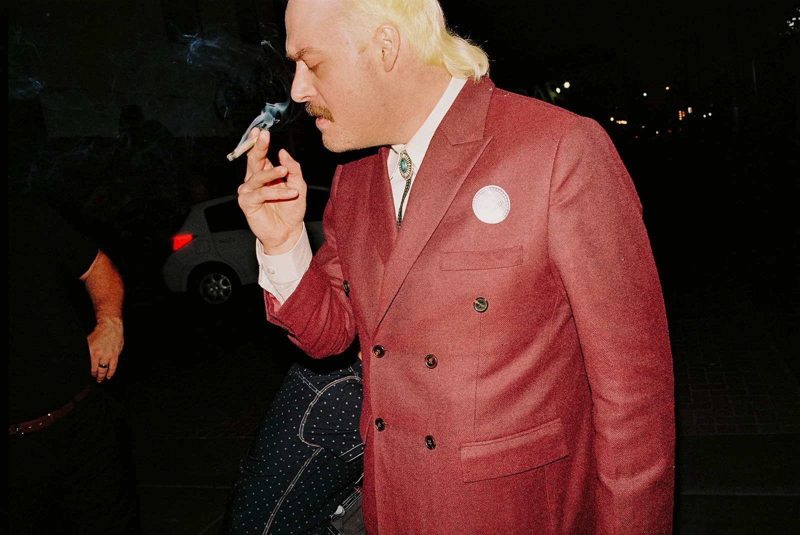 A man with blond hair and a mustache, dressed in a red double-breasted suit, is smoking a cigarette at night.