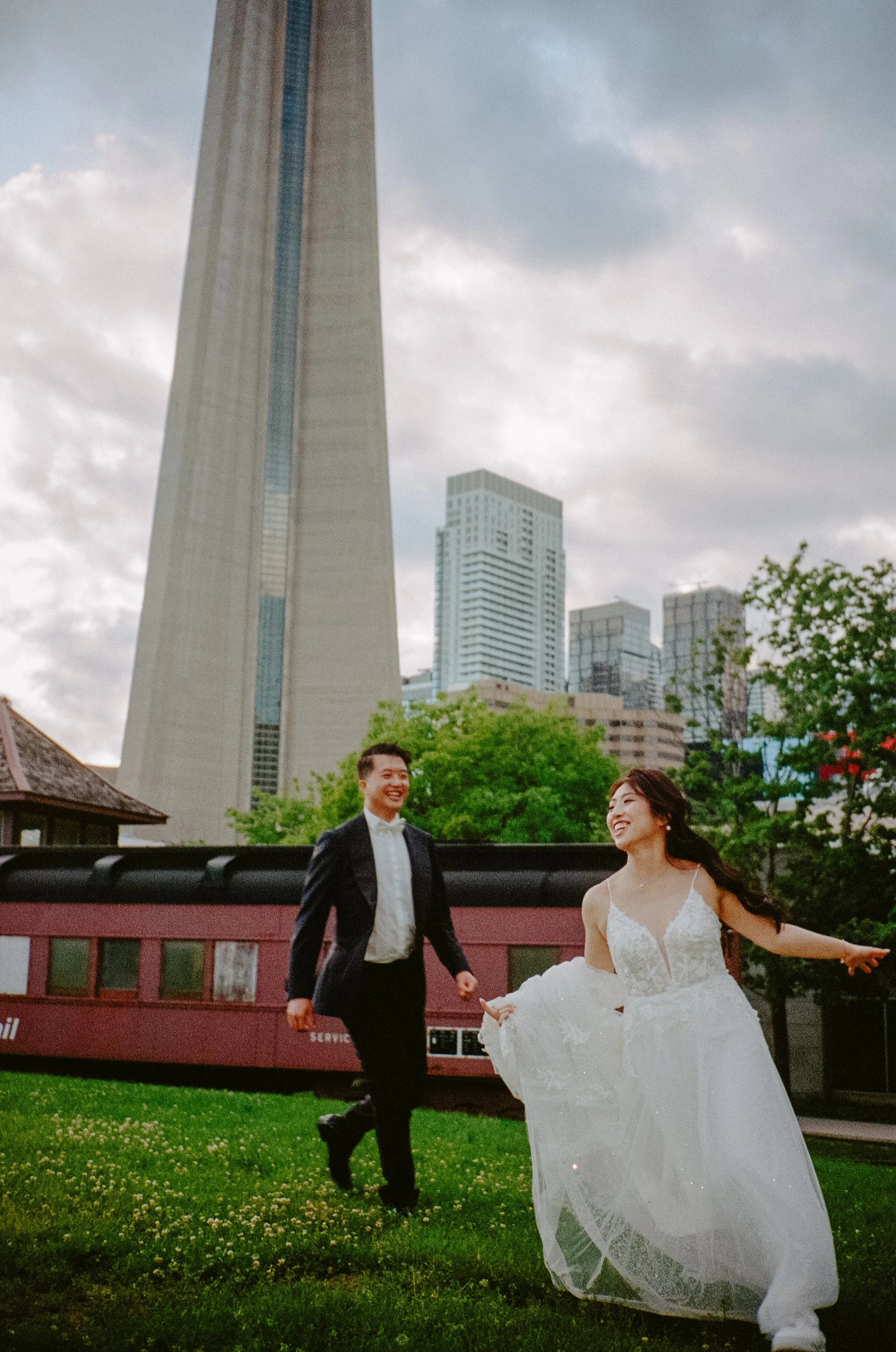 A couple in wedding attire running on grass with city skyscrapers in the background.