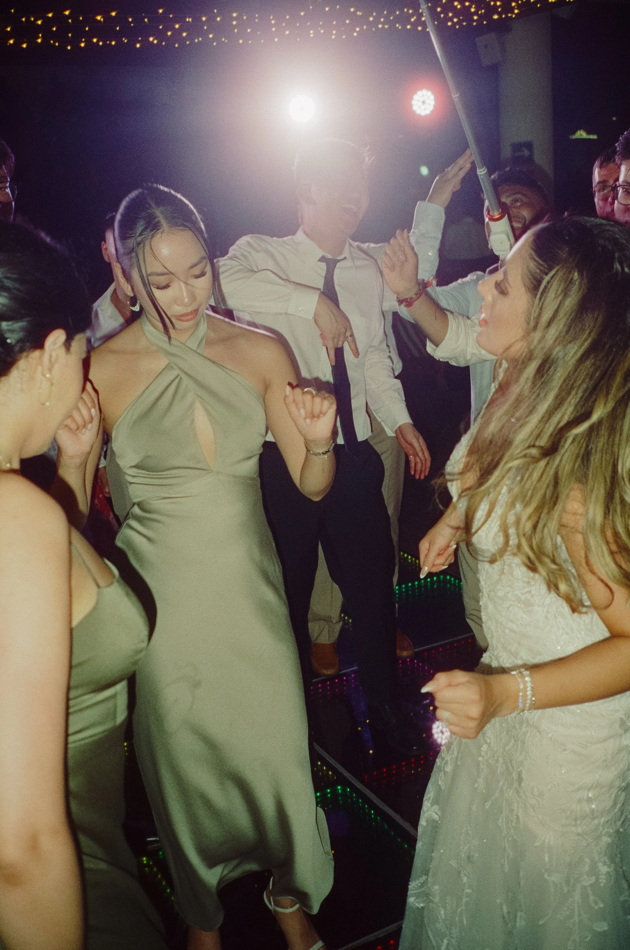 People dancing at a party or celebration with colorful lighting and a DJ or music equipment in the background.