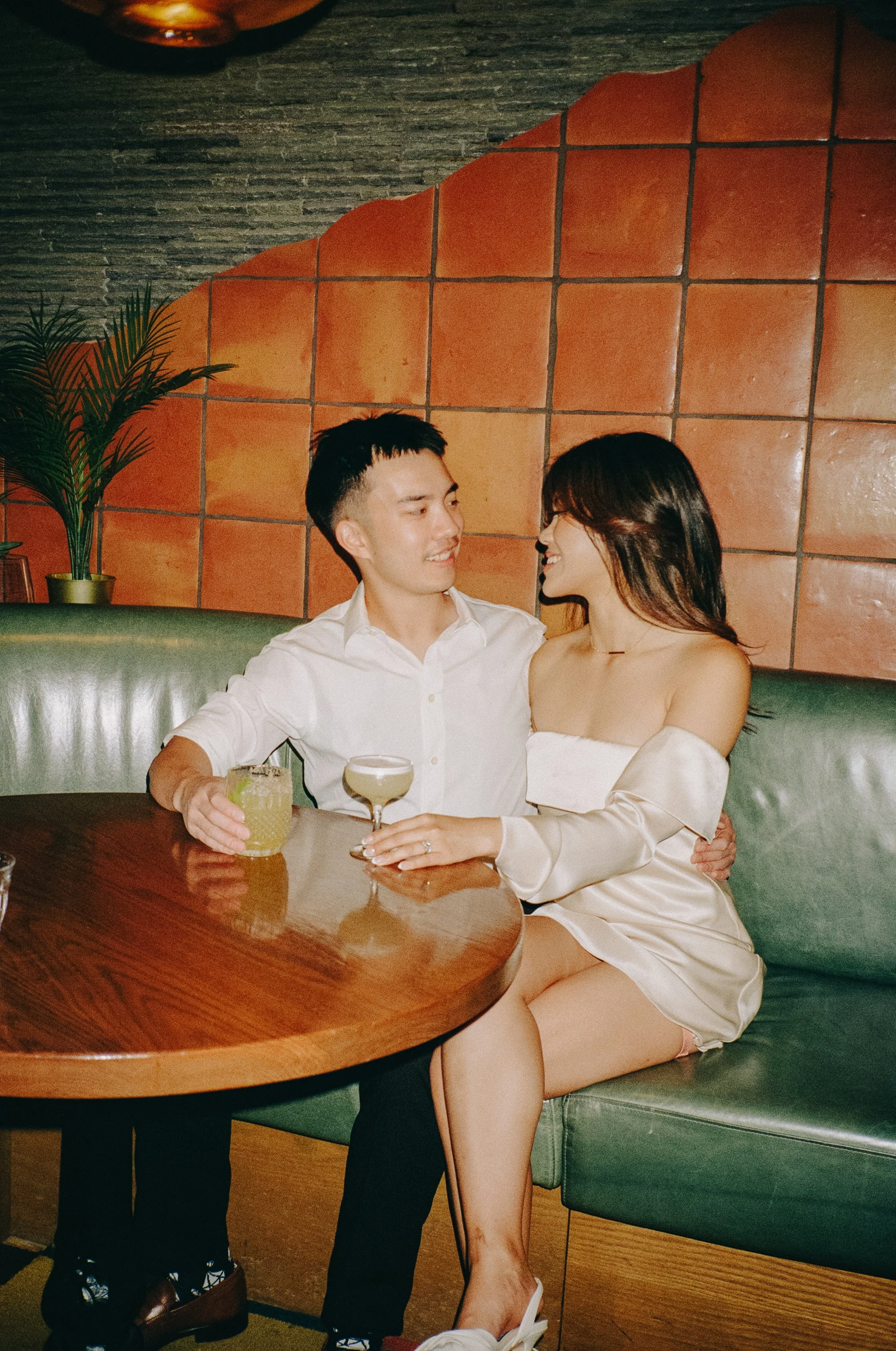 A young man and woman sitting on a green booth bench at a restaurant or bar, sharing drinks and looking at each other affectionately. The man is holding a yellow cocktail, and the woman has a champagne glass.