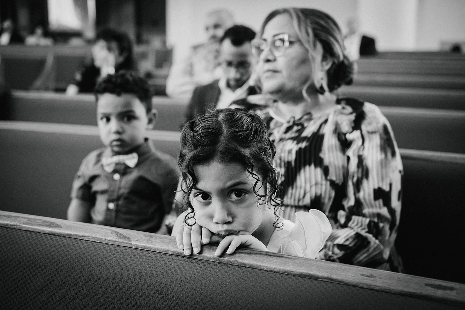 Black and white photo of a young girl with curly hair leaning on a church pew, looking directly at the camera with her head resting on her hands. Behind her, a woman with glasses and a striped blouse looks to the side, and a young boy with a bow tie 