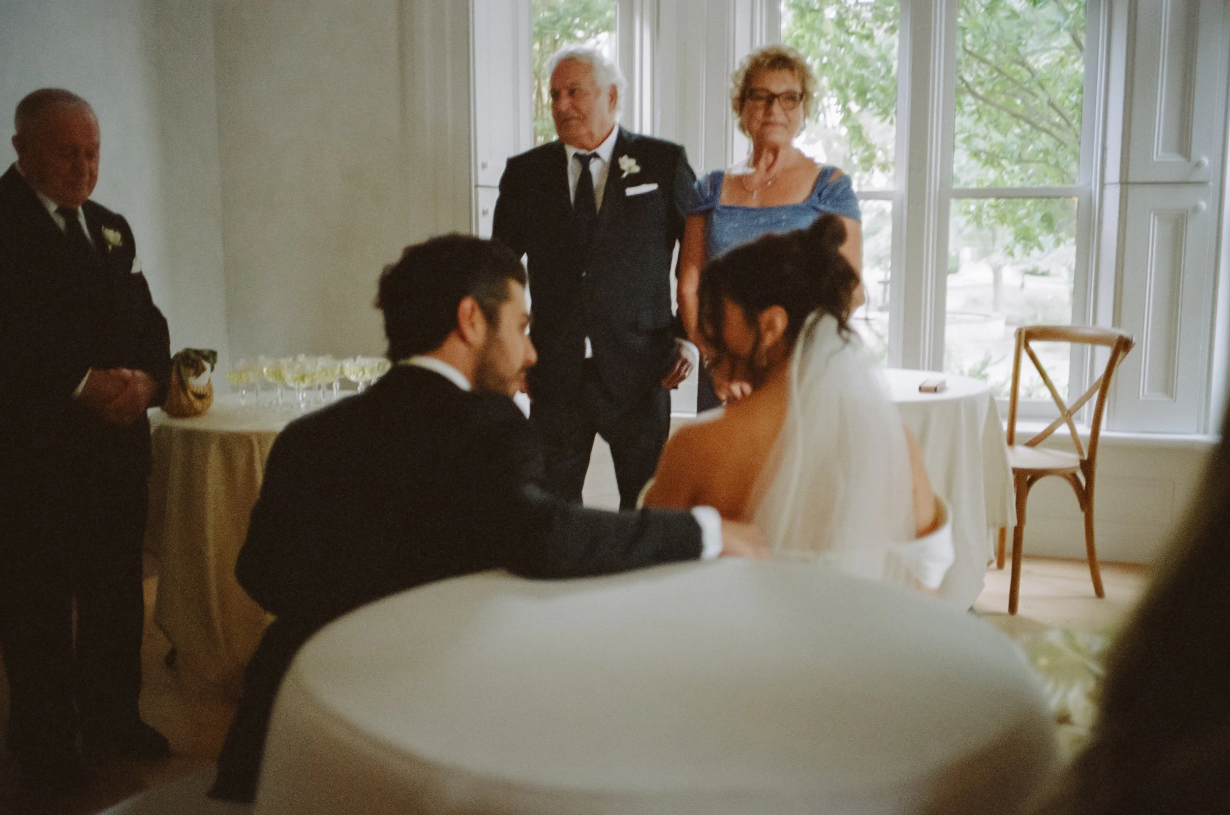 A wedding reception scene where a bride and groom are seated at a table, with the groom holding the bride's hand. Three older seated guests and two standing guests are observing them near large windows in a bright room.