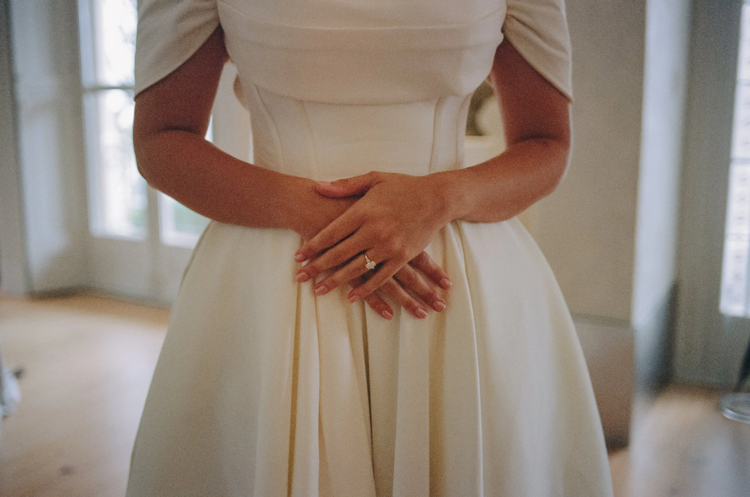 Woman in an elegant cream dress with her hands clasped together at waist, showing a wedding ring, in a softly lit room with large windows in background.