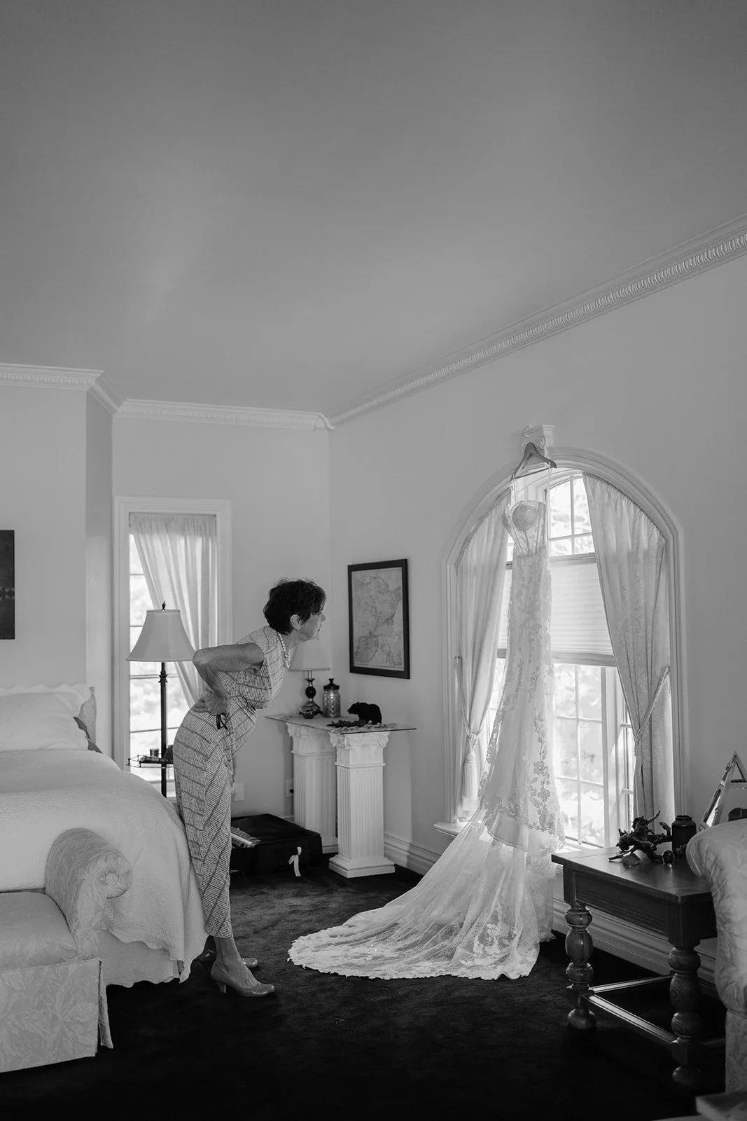 A woman looks at a wedding dress hanging by a window in a bedroom.