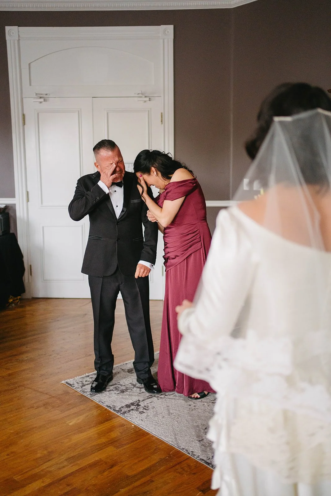 A man in a black tuxedo and a woman in a burgundy dress are emotional, touching foreheads, crying, and smiling in a room with wooden floors, a small rug, and a white closed door. Another woman in a white wedding dress with a veil is partially visible