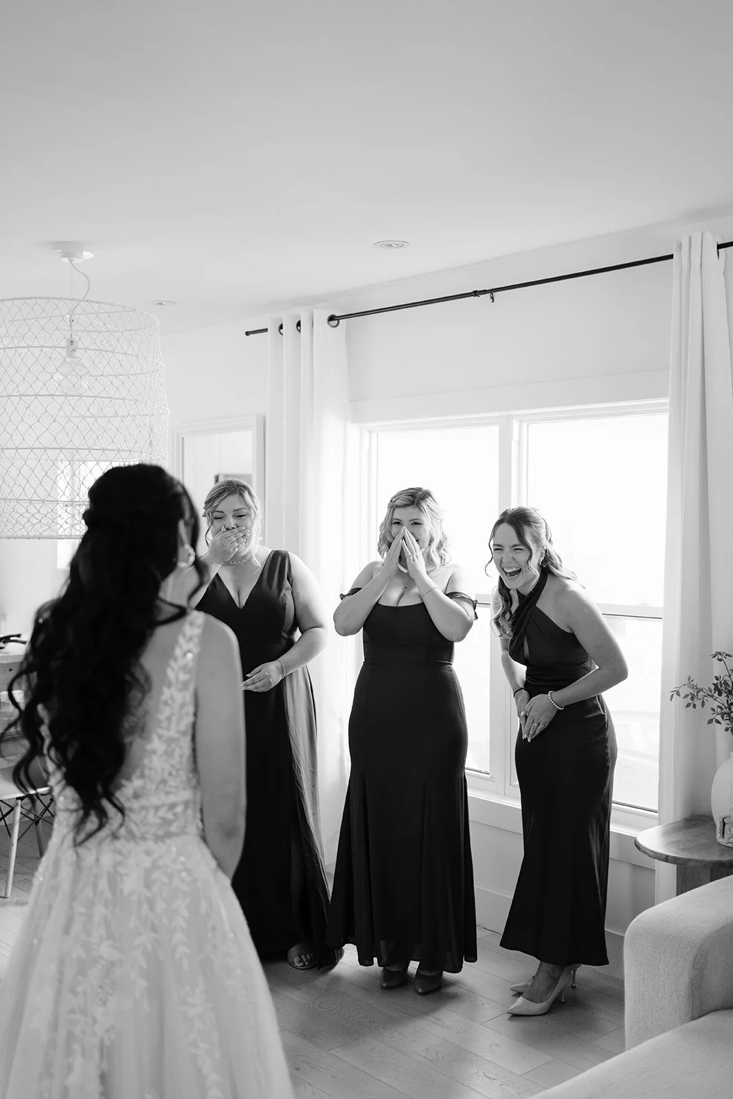 Four women in formal dresses inside a bright room with large windows, one woman facing away in a white wedding gown, three women reacting emotionally, possibly at a wedding or special event.