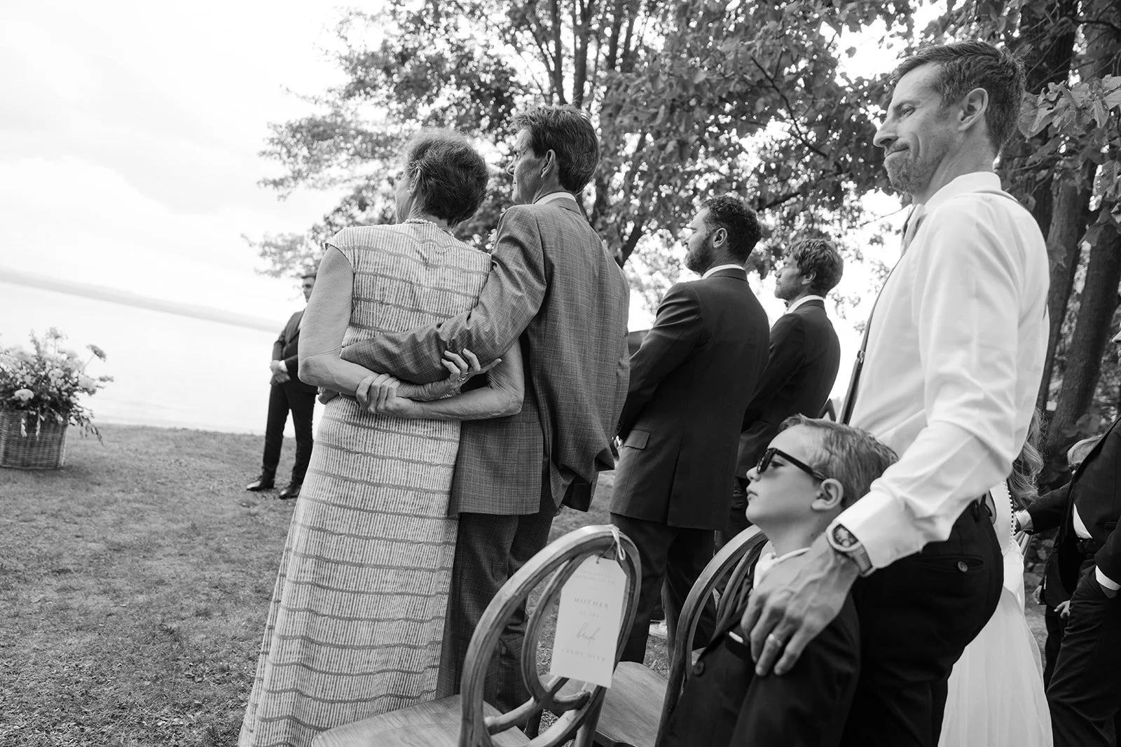 People attending an outdoor wedding ceremony, standing together with some holding each other, during the daytime with trees in the background.