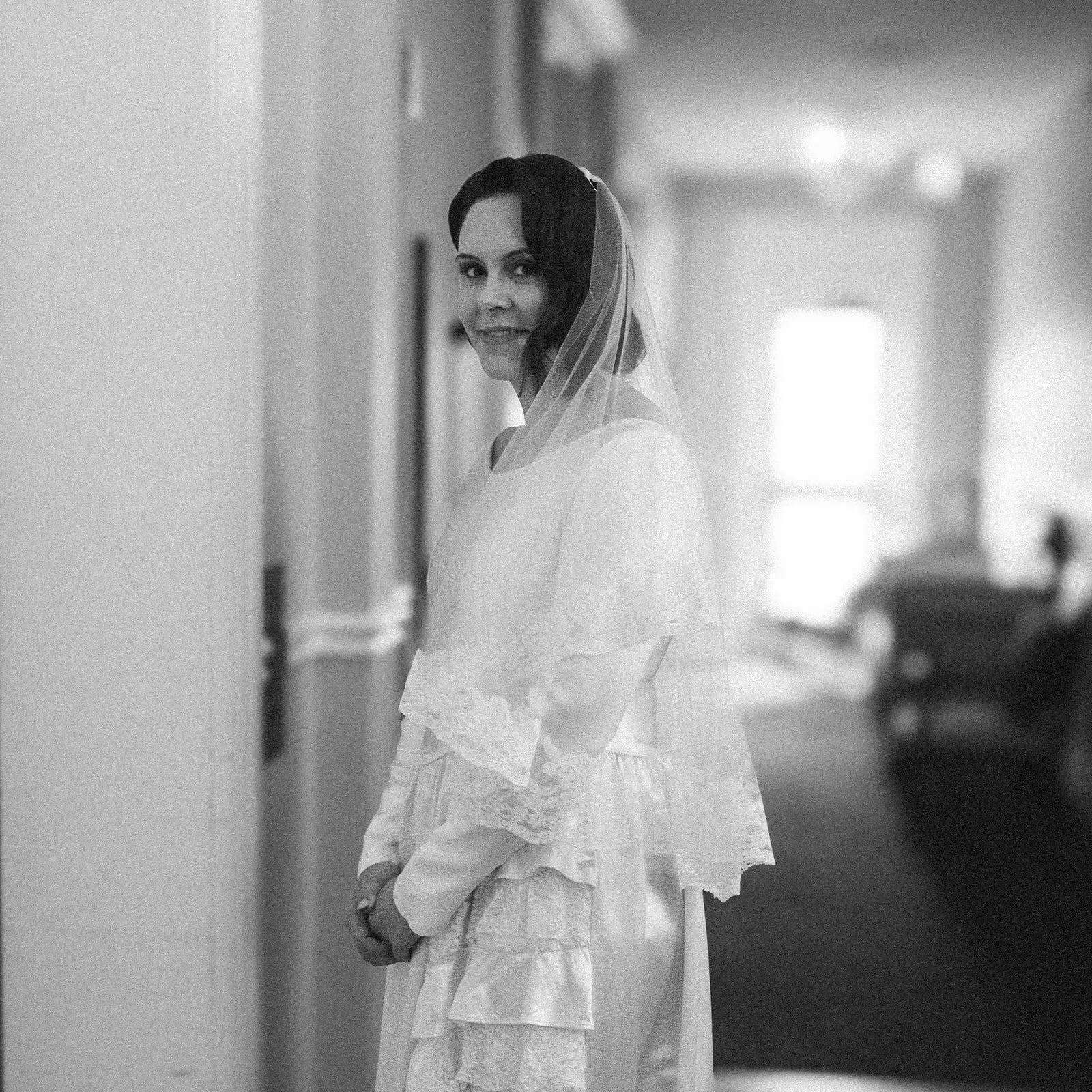 Black and white photo of a woman in a bridal gown with a veil, standing in a hallway, smiling at the camera.
