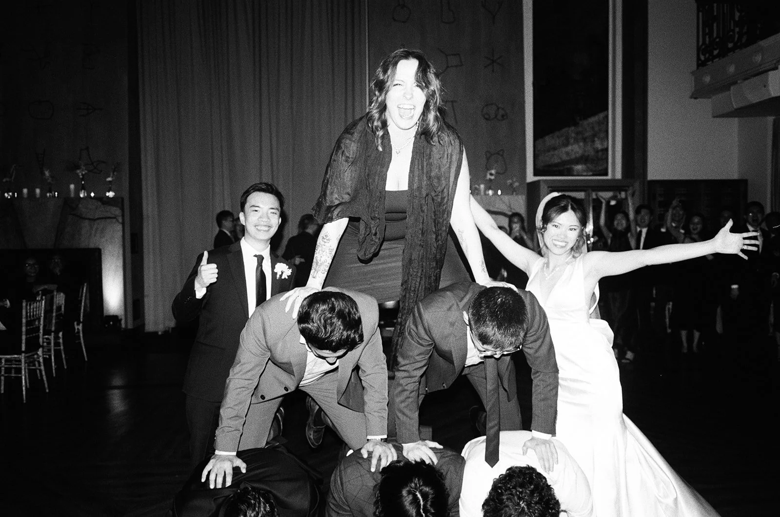 A group of people forming a human pyramid at a celebration, with a woman at the top smiling and others supporting her underneath in a festive setting.