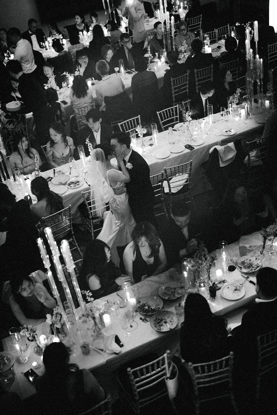 A black-and-white photo of a wedding reception with guests seated at long decorated tables, plates of food, candles, and floral centerpieces. A bride and groom are dancing in the center of the room.