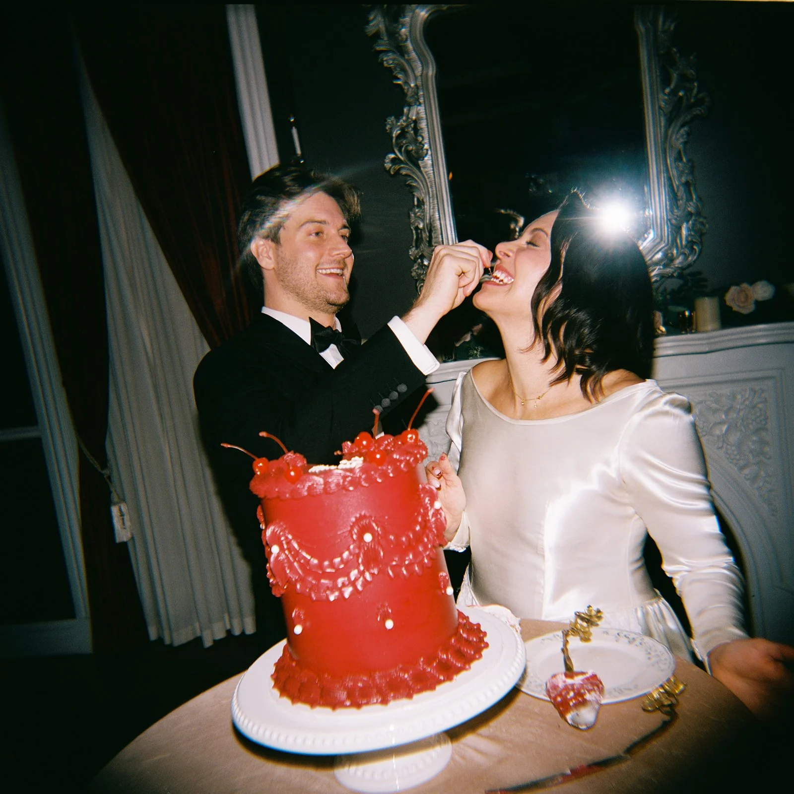 A man in a tuxedo feeds a piece of cake to a woman in a white dress during a celebration, with a red cake topped with cherries and decorative frosting on the table.