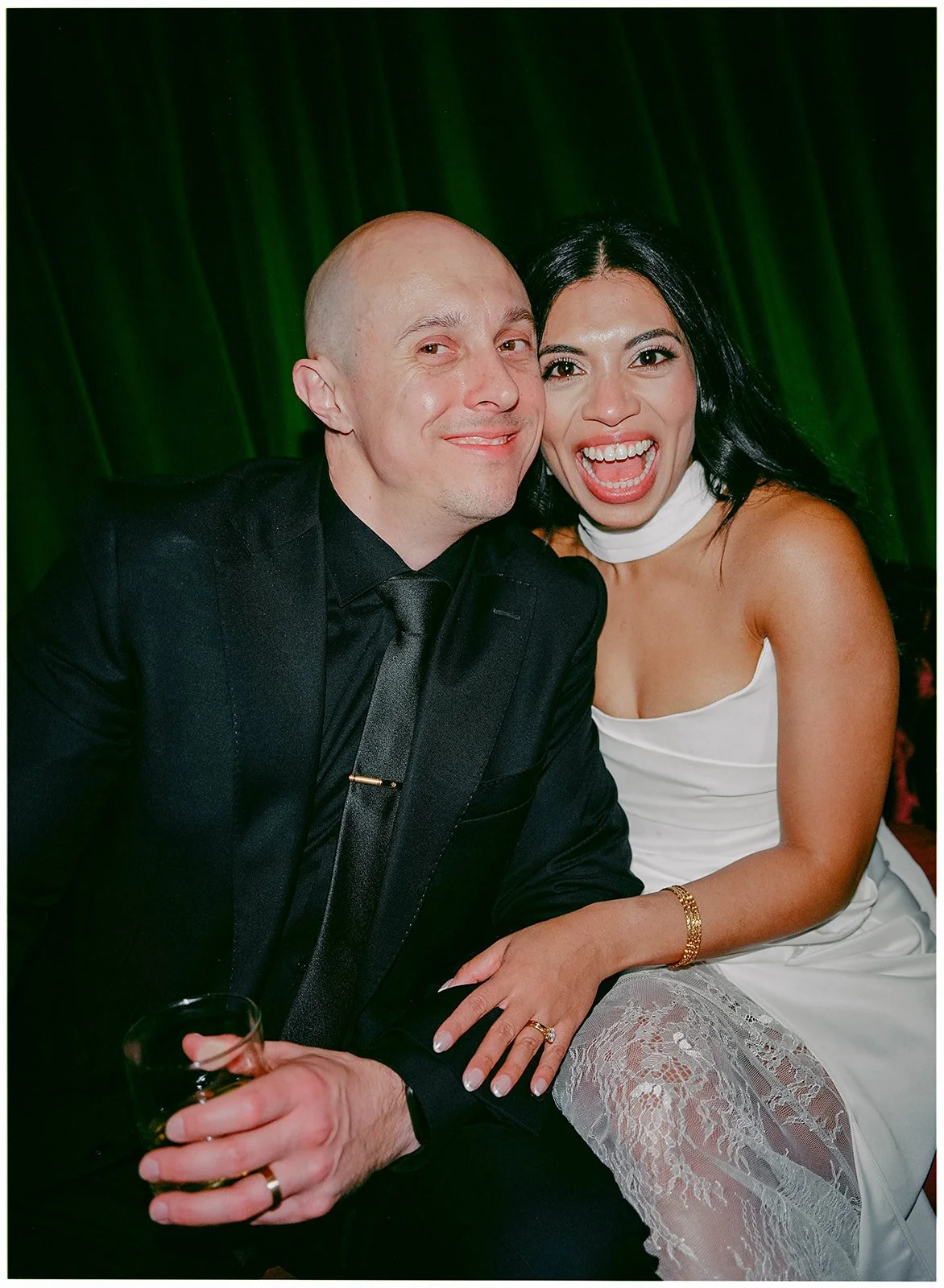 A man and woman sitting close together, smiling and posing for a photo in front of a green curtain. The man is holding a drink, wearing a black suit and tie, and has a bald head. The woman has long black hair, is wearing a white dress with lace detai