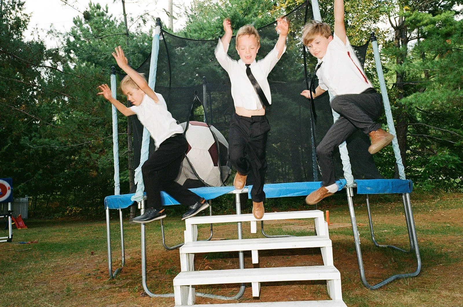 Four young boys in dress shirts and pants jumping on a trampoline outdoors with trees in the background.