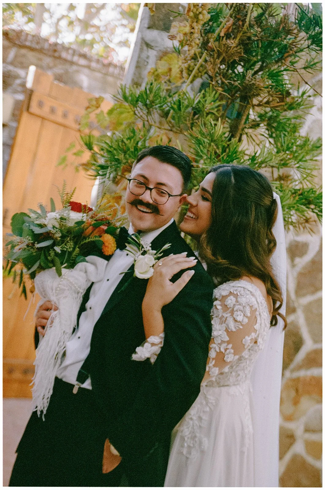 A bride and groom smiling joyfully at each other, with the bride touching the groom's shoulder and holding a bouquet, dressed in wedding attire, standing in front of a leafy background.