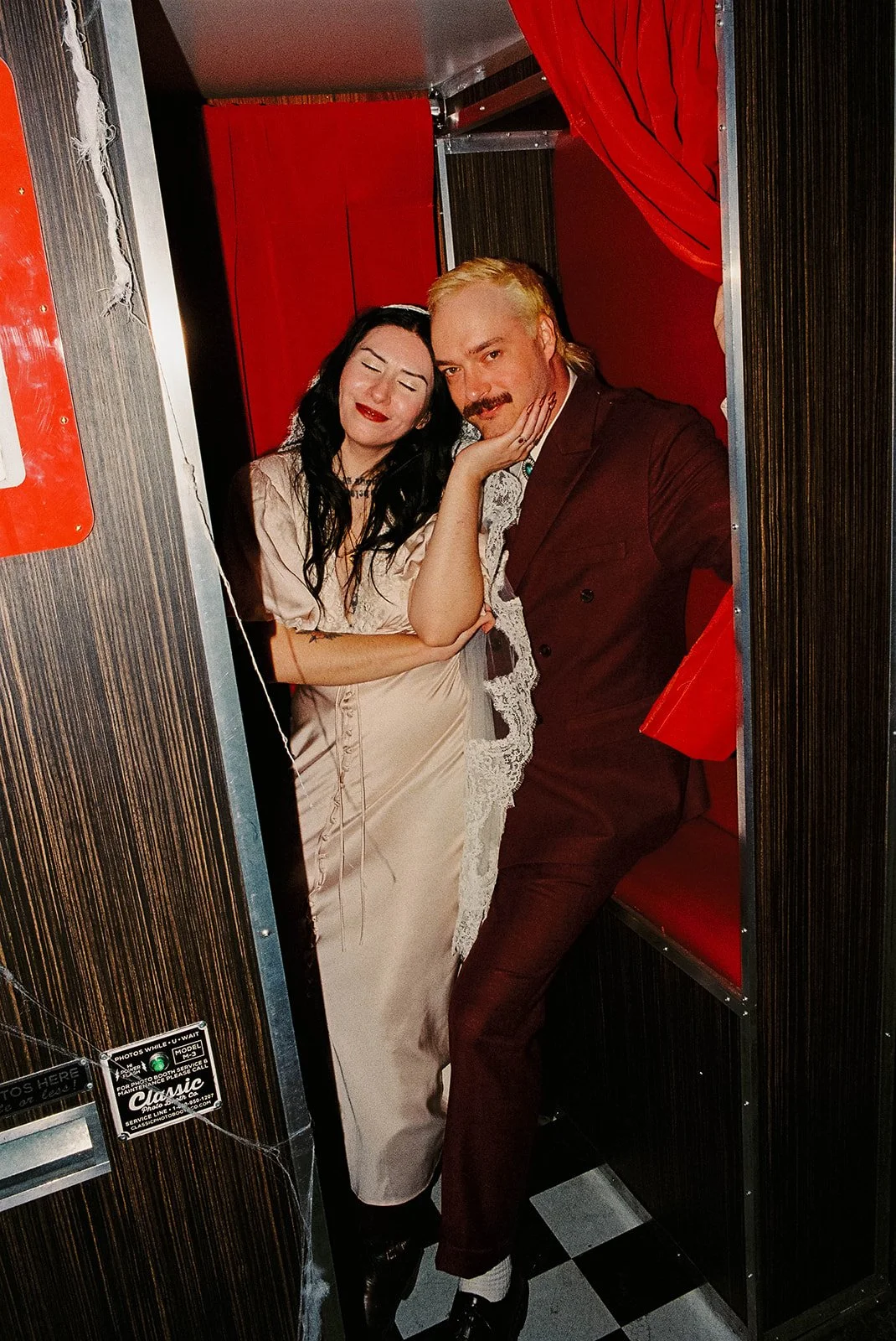 Two people, a woman with dark hair and a man with blond hair and a mustache, posing inside a small red booth. The woman is wearing a light beige satin dress and the man is dressed in a burgundy suit. They are affectionately leaning against each other