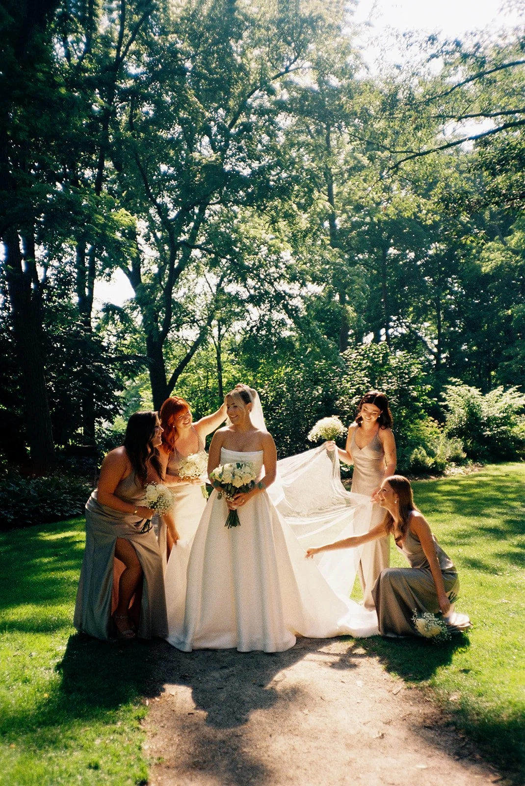 A bride in a white wedding dress holding a bouquet of flowers, surrounded by five bridesmaids in beige dresses, some holding bouquets, in a lush green garden with tall trees and sunlight streaming through.