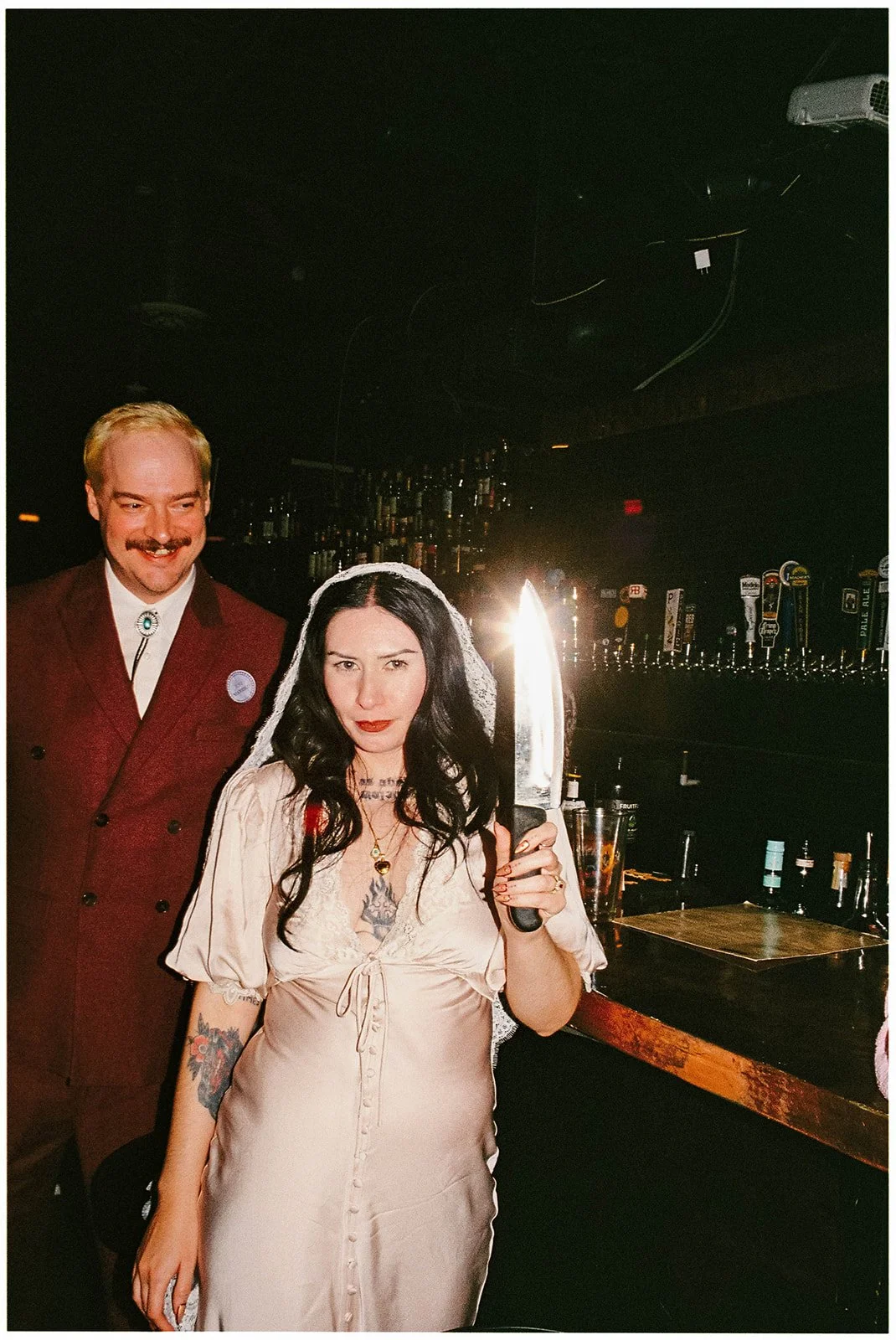 A woman with long black hair holding a large knife while wearing a beige satin dress and standing in a bar, with a man in a maroon suit standing behind her, smiling.