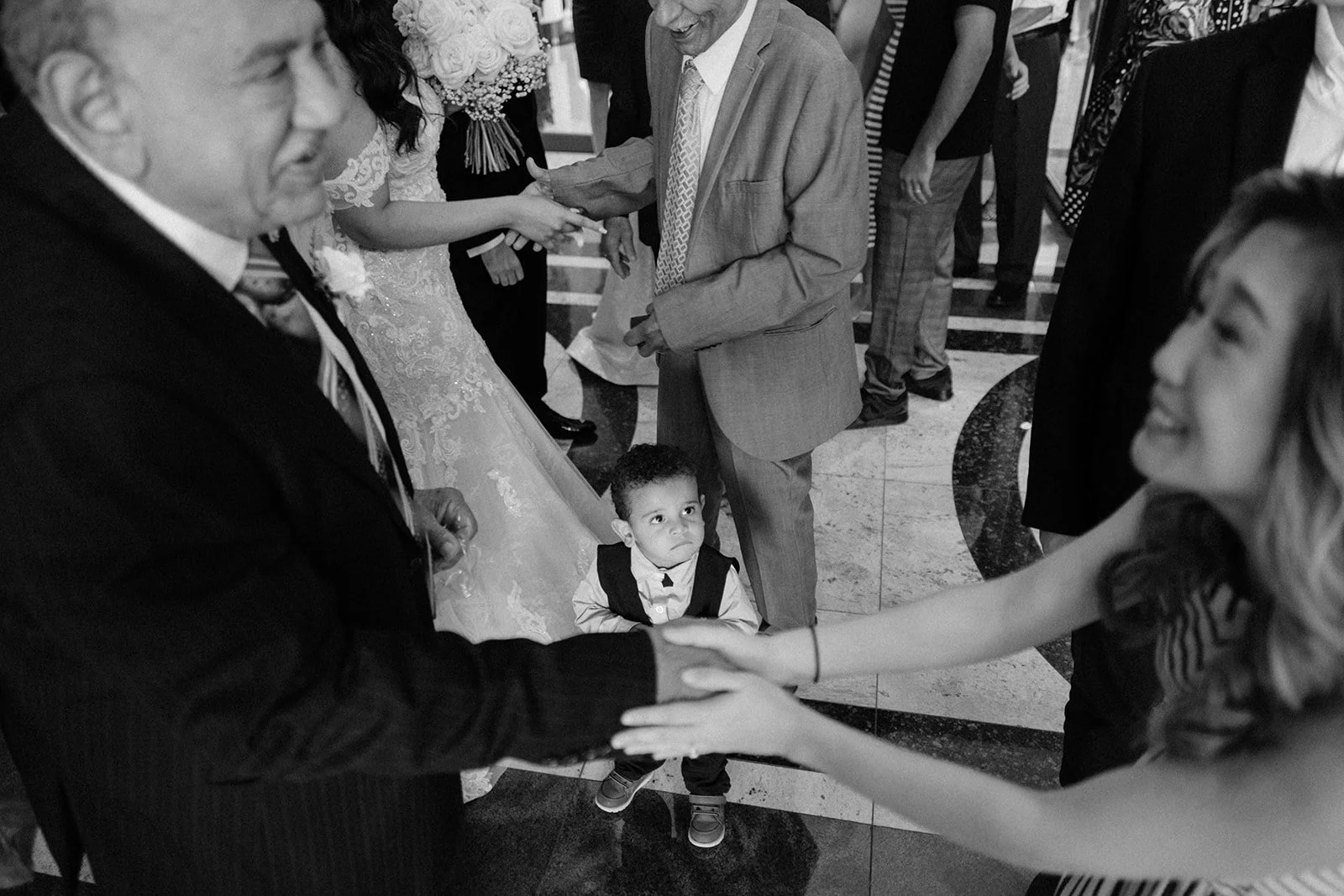 A young boy looking up while holding hands with a woman, at a formal gathering or celebration with people dancing and socializing.