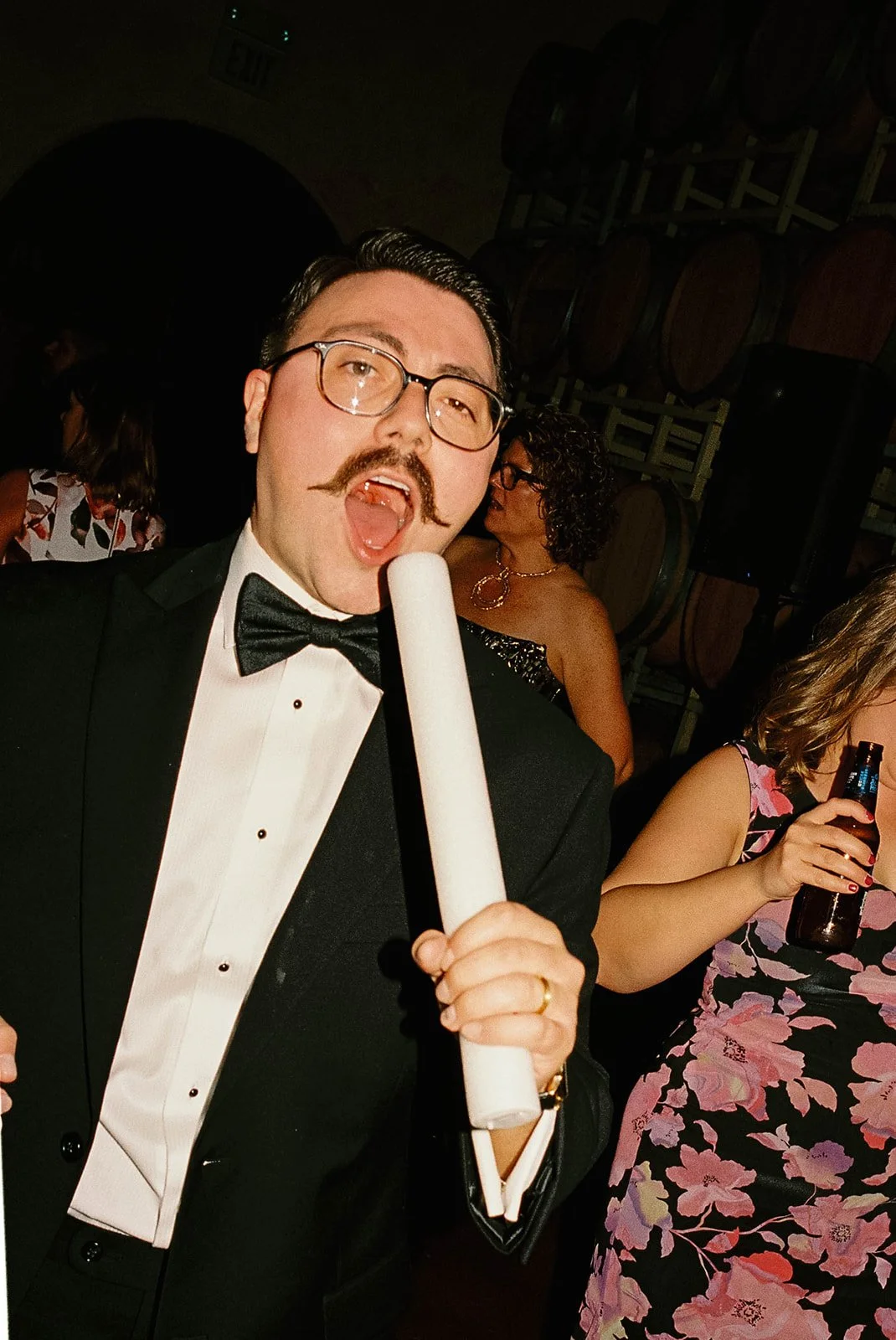 A man in a tuxedo with glasses and a mustache singing into a microphone at a party.