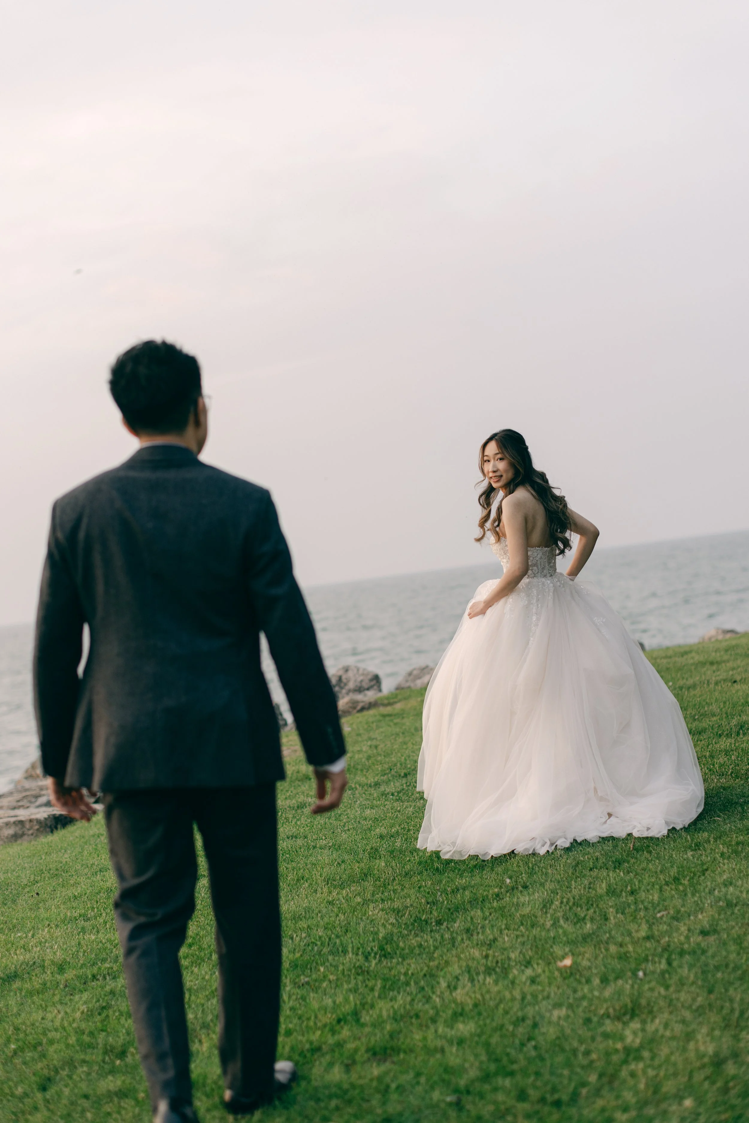A bride in a white wedding gown smiling at a man in a suit on a grassy area near the water.