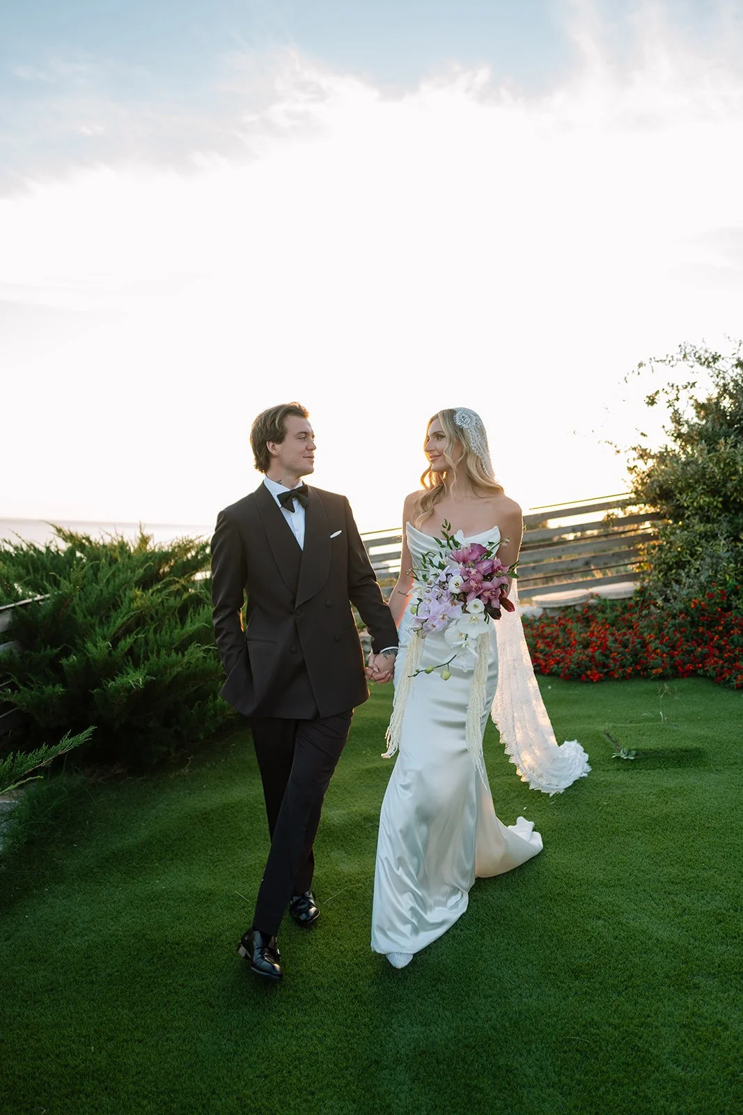 A bride and groom holding hands outside at sunset, walking on grass, with greenery and flowers in the background.