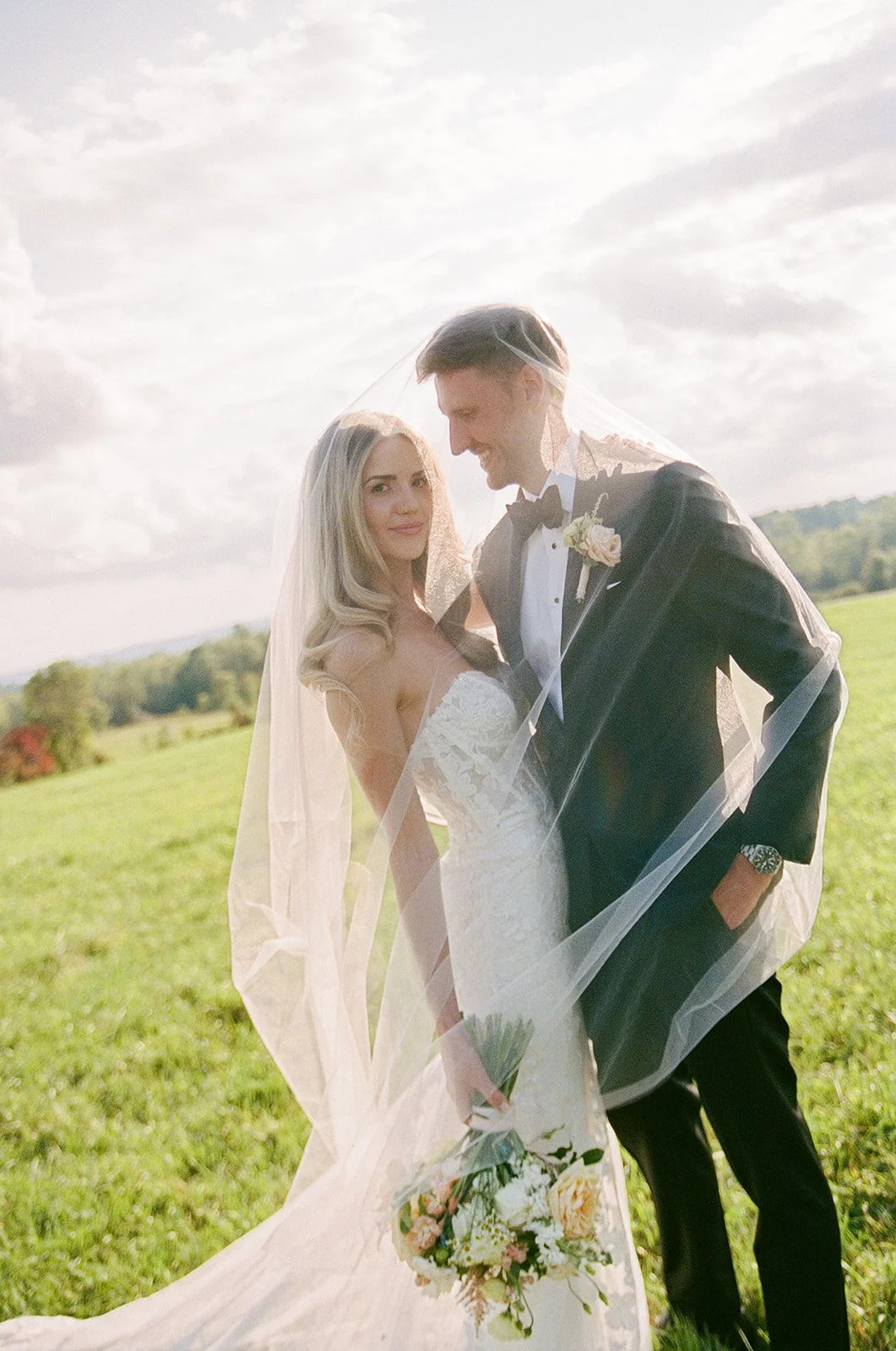 A bride and groom in wedding attire standing outdoors on a grassy field, with a veil draped over them and a bouquet of flowers, under a cloudy sky.