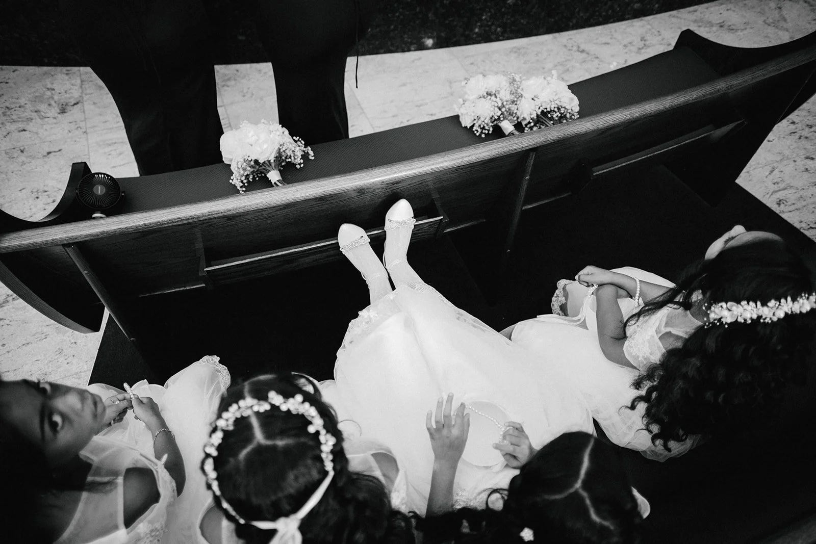 Top-down view of a girl in a white dress and tiara lying on a church pew with three other girls nearby, bouquets on the pew, and a person standing at the end of the pew.