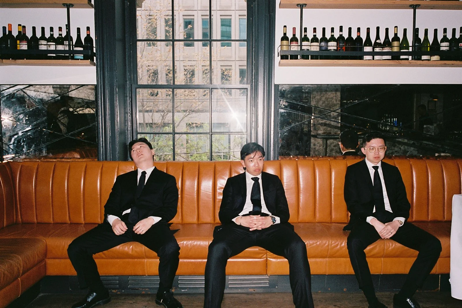 Three men dressed in black suits and ties sitting on a leather booth in a restaurant or bar, with large windows behind them and bottles of wine on shelves above.