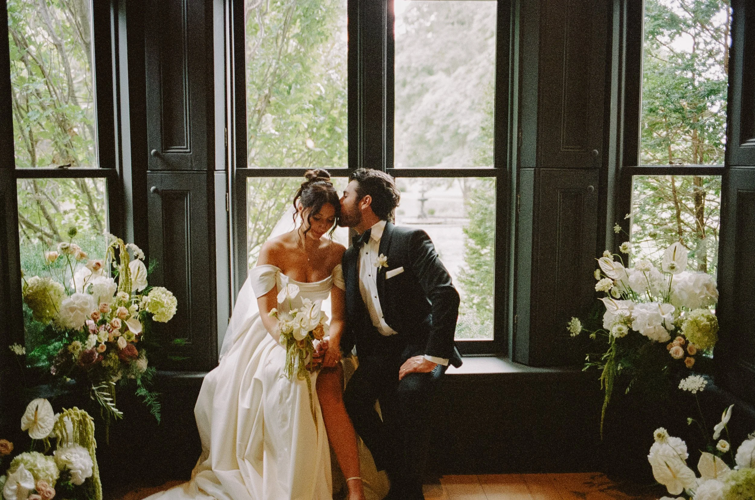 A bride and groom in wedding attire sitting close together near a large window with greenery outside, surrounded by floral arrangements.