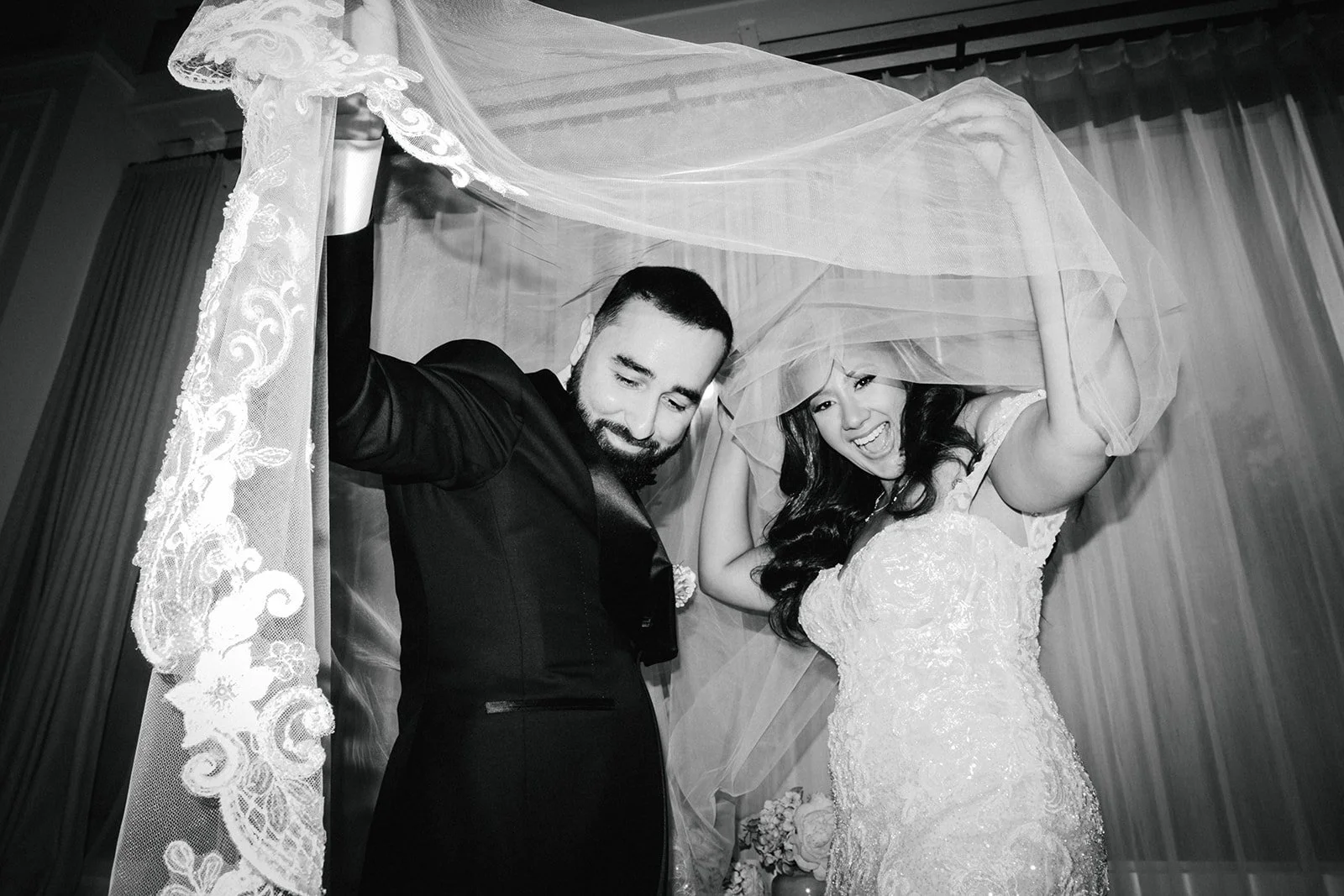 A newlywed couple standing under a wedding canopy, with the groom in a black tuxedo and the bride in a white wedding dress, both smiling and holding the veil over their heads.