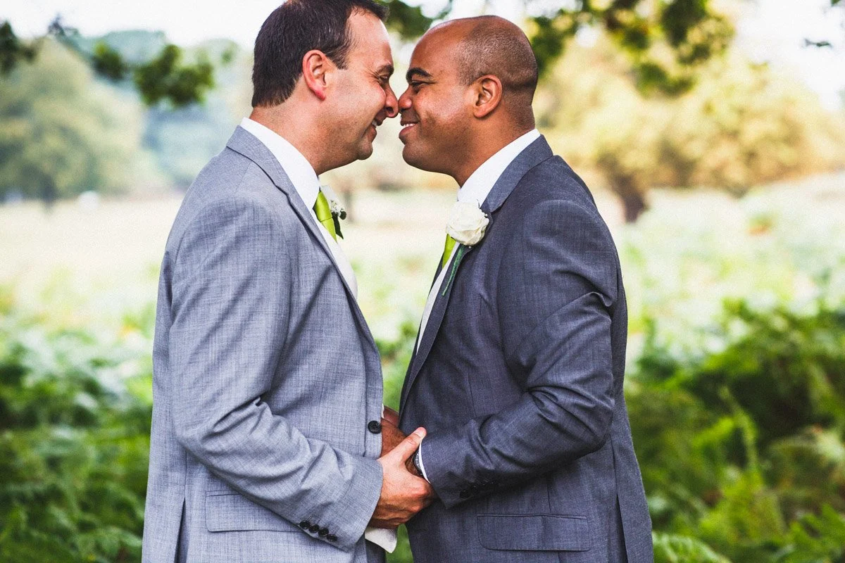 Two men in wedding suits embracing and touching foreheads outdoors during daytime.