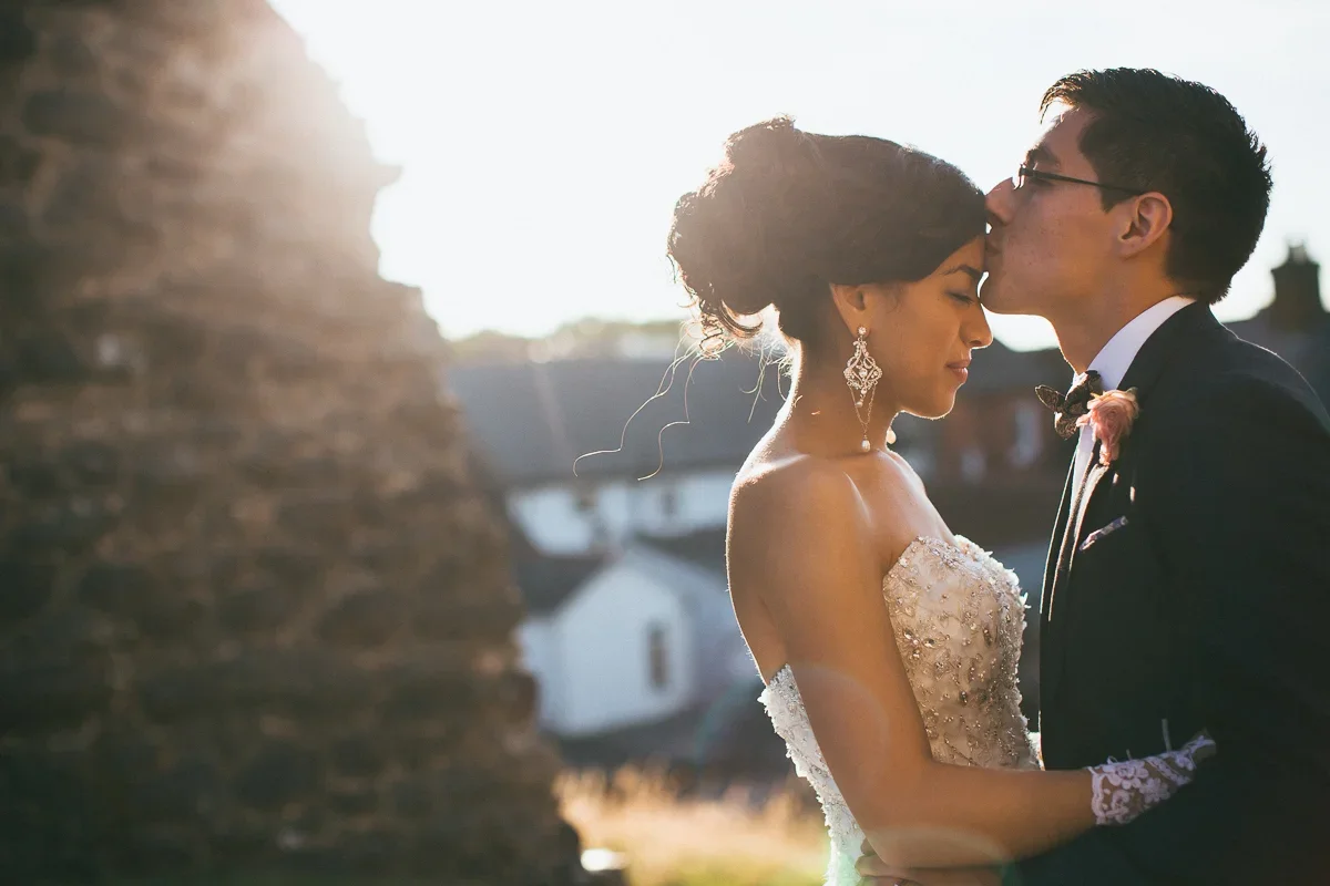 A bride and groom sharing a kiss on their wedding day in a romantic outdoor setting with sunlight.