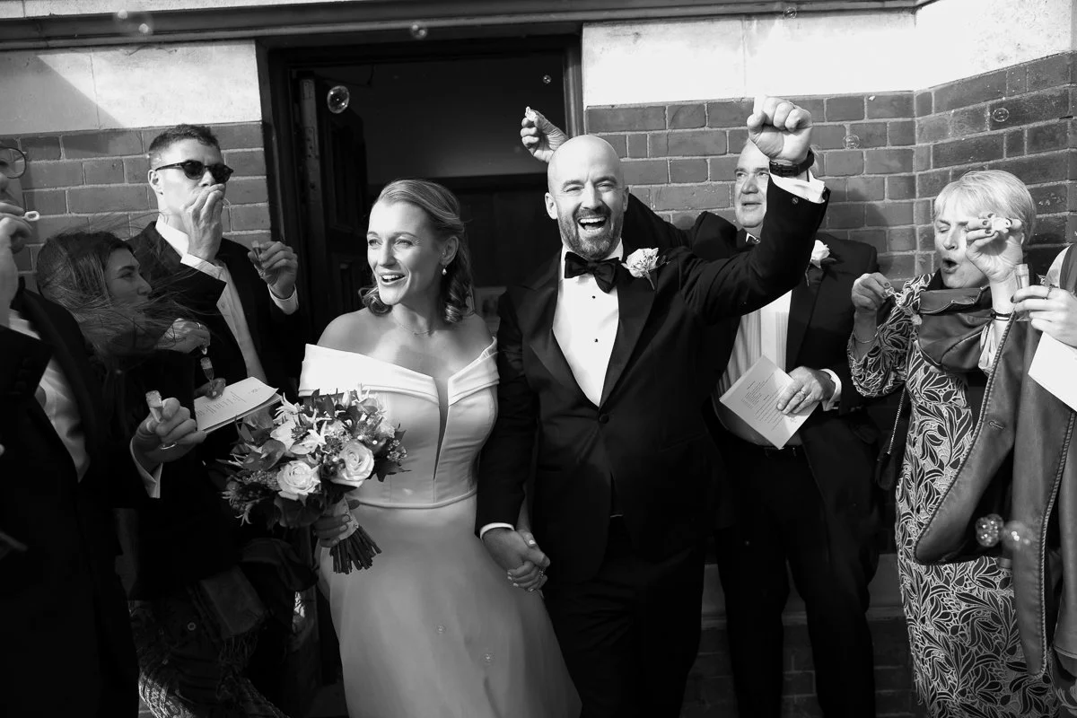 A wedding celebration with a bride and groom holding hands, smiling, surrounded by guests celebrating and blowing bubbles.