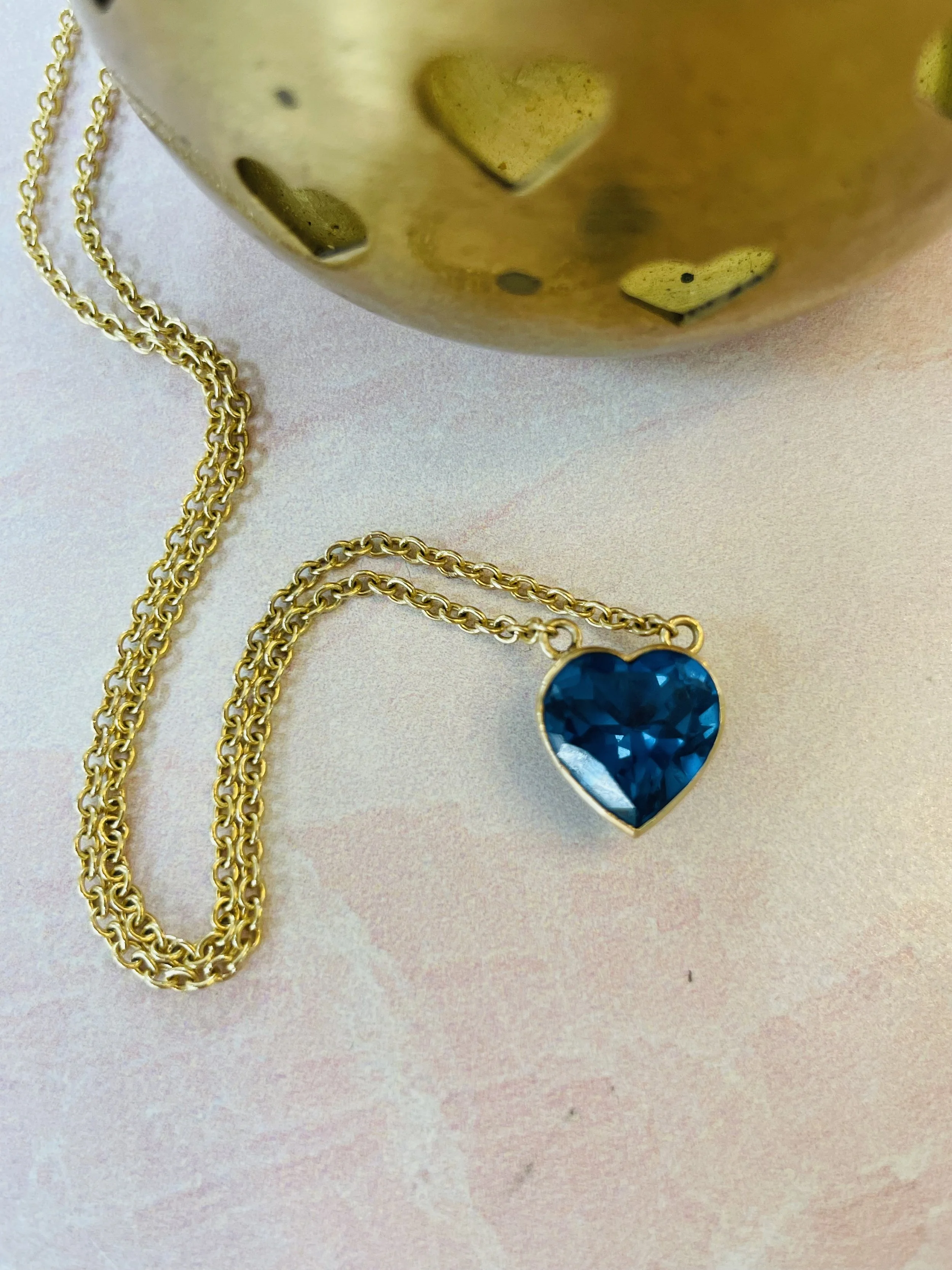 Gold chain necklace with a heart-shaped blue gemstone pendant, placed on a pink surface next to a golden decorative object with heart-shaped cutouts.