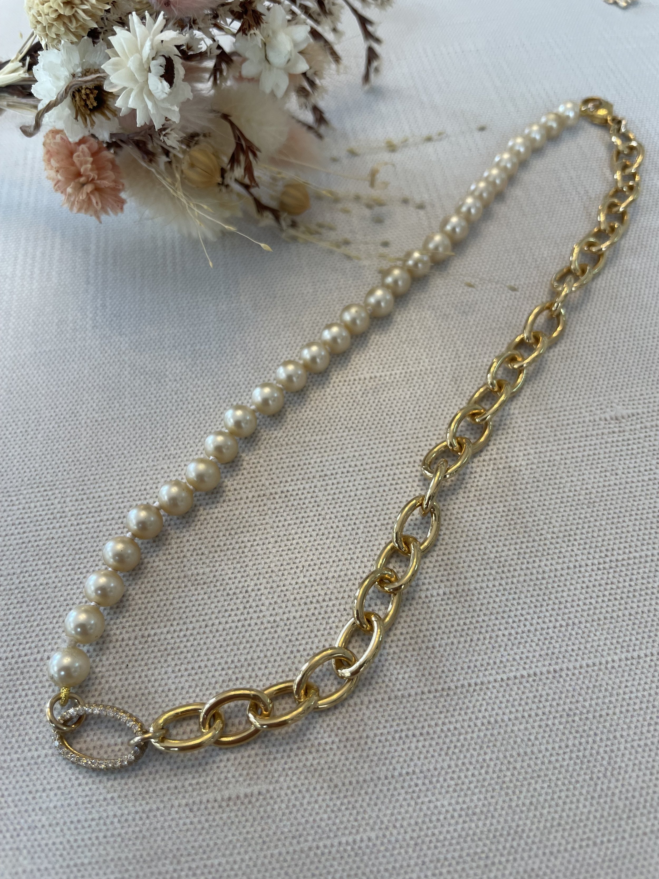 A gold chain necklace and a string of white pearls arranged on a textured surface with a dried flower bouquet nearby.