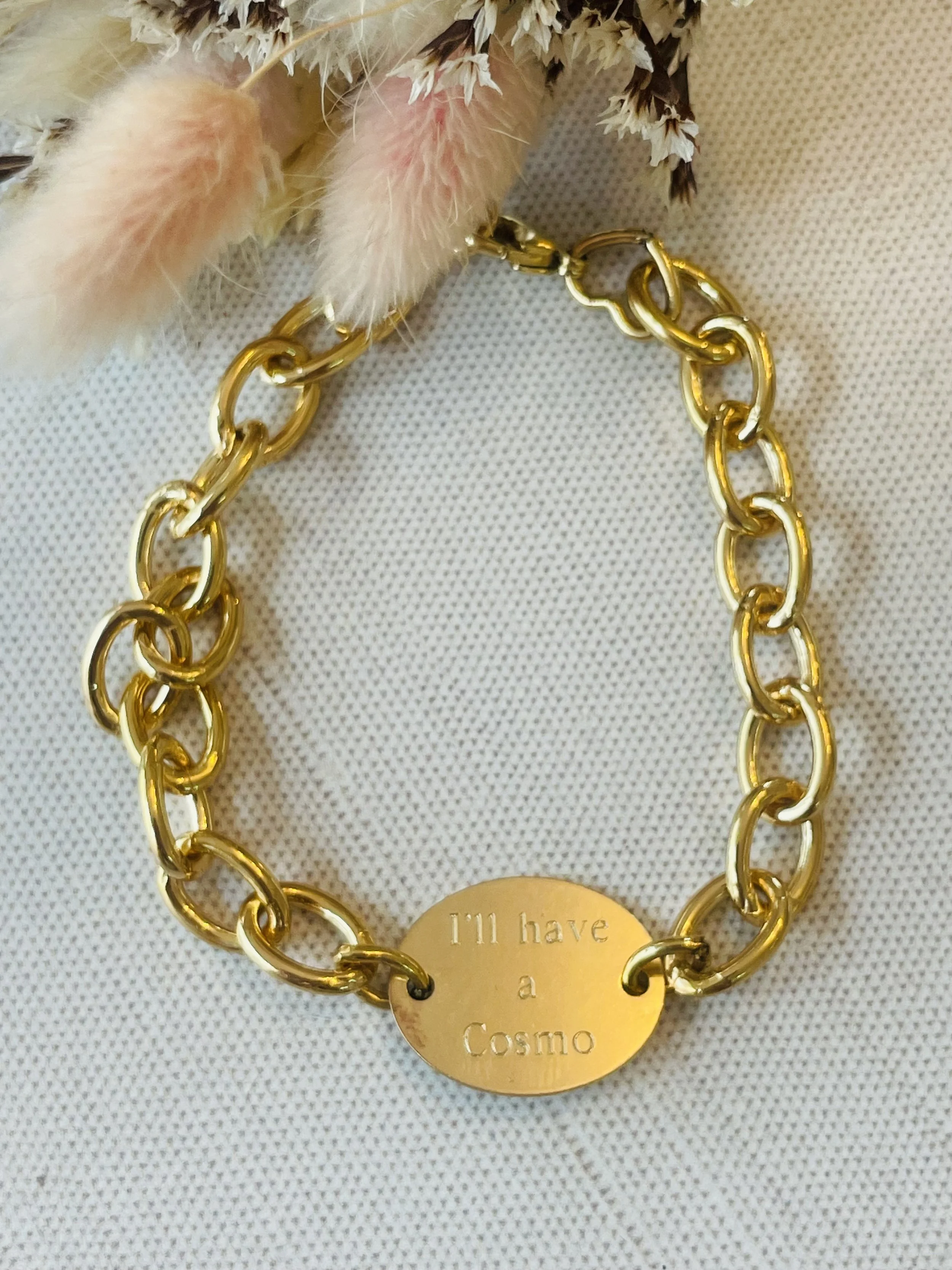 Gold chain bracelet with an oval engraved charm that says "I'll have a Cosmo," placed on a textured white surface, accompanied by dried flowers or foliage.
