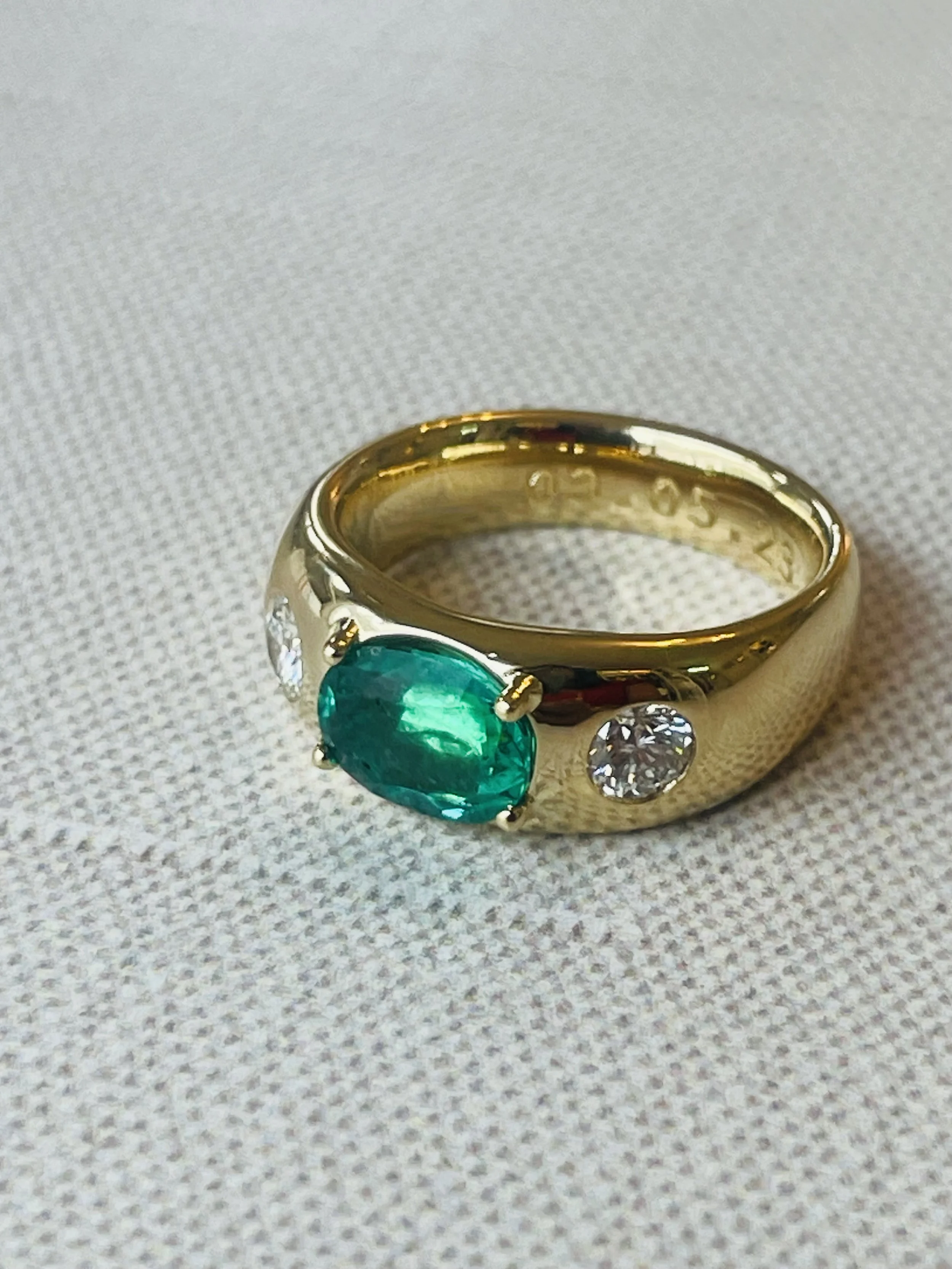 Gold ring with an oval emerald gemstone flanked by two clear diamonds on a white textured surface.