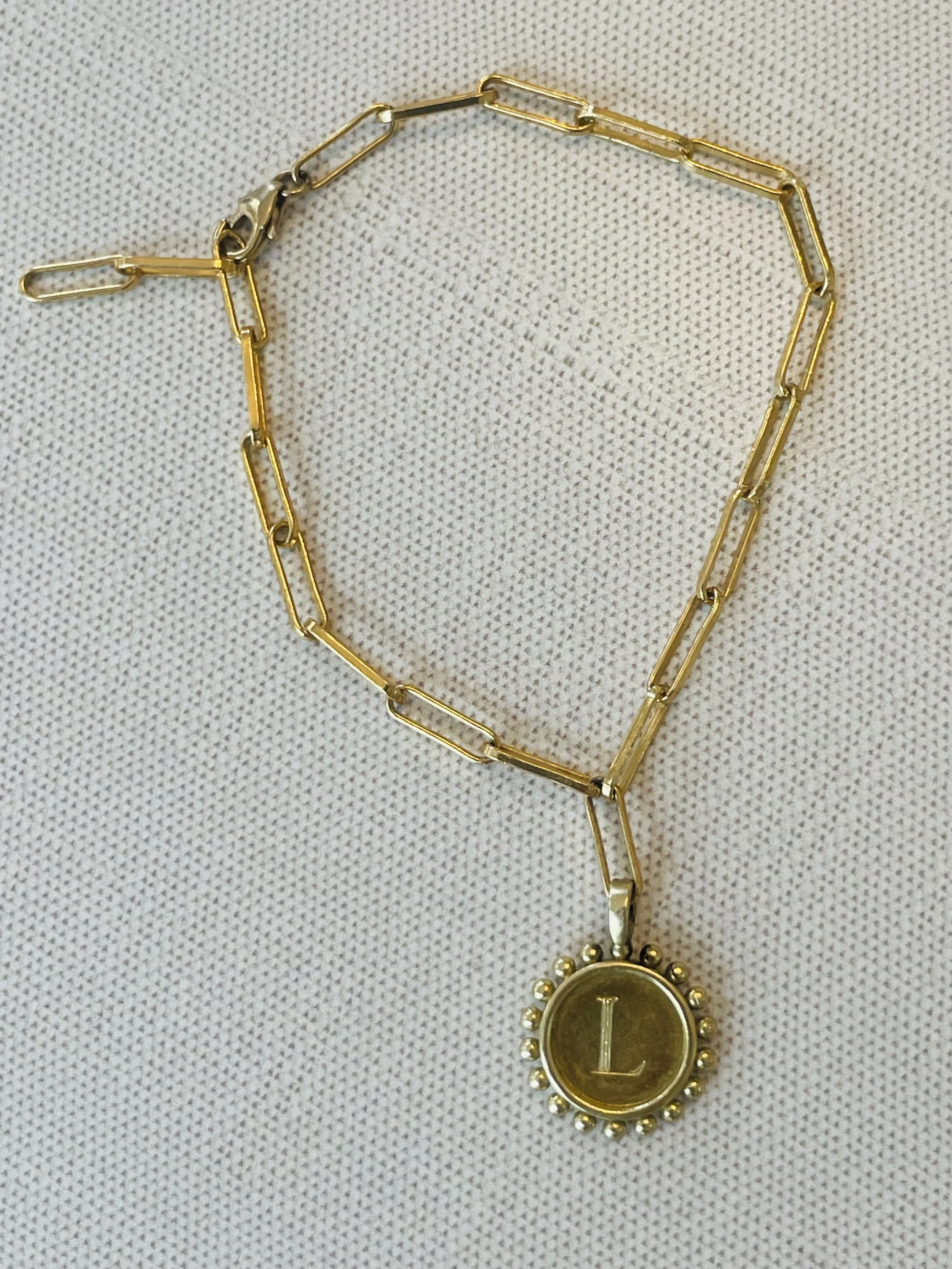 Gold necklace with rectangular links and a round pendant with an 'L' engraved, surrounded by small beads, on a light fabric background.