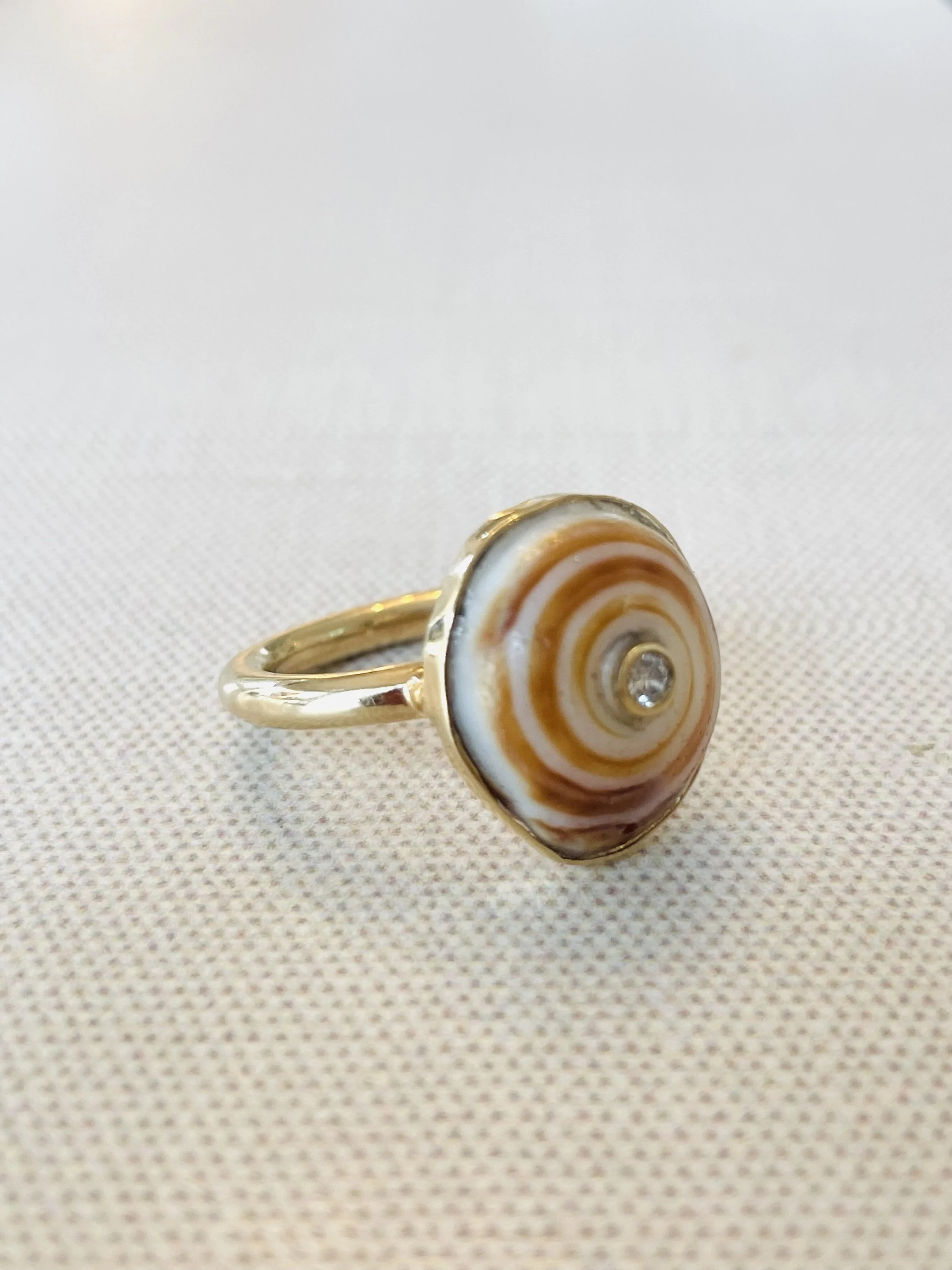 Gold ring with a spiral shell design and a small central gemstone placed on a textured beige surface.