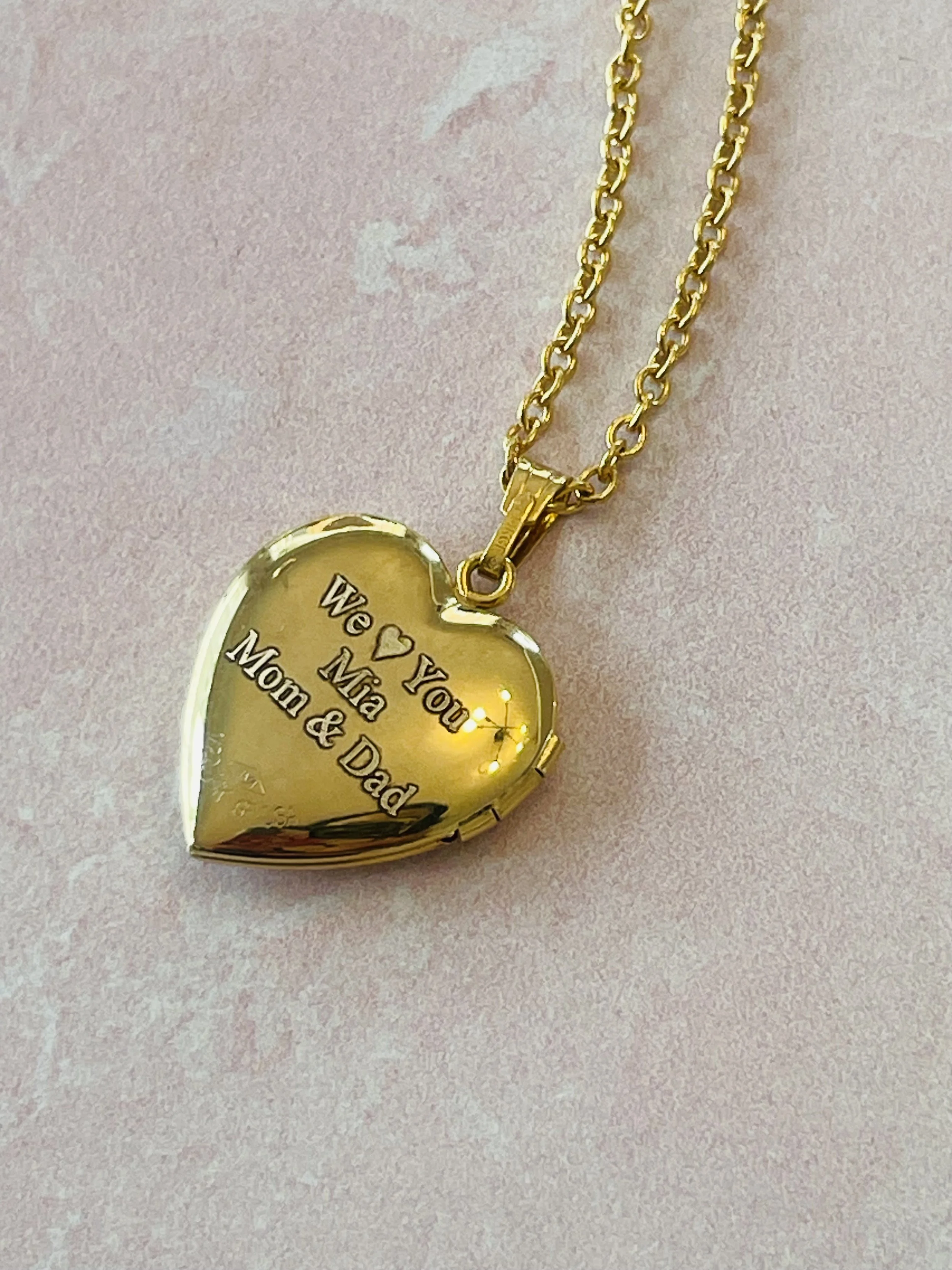 Gold heart-shaped pendant necklace with engraved message for Mom & Dad against a pink background.