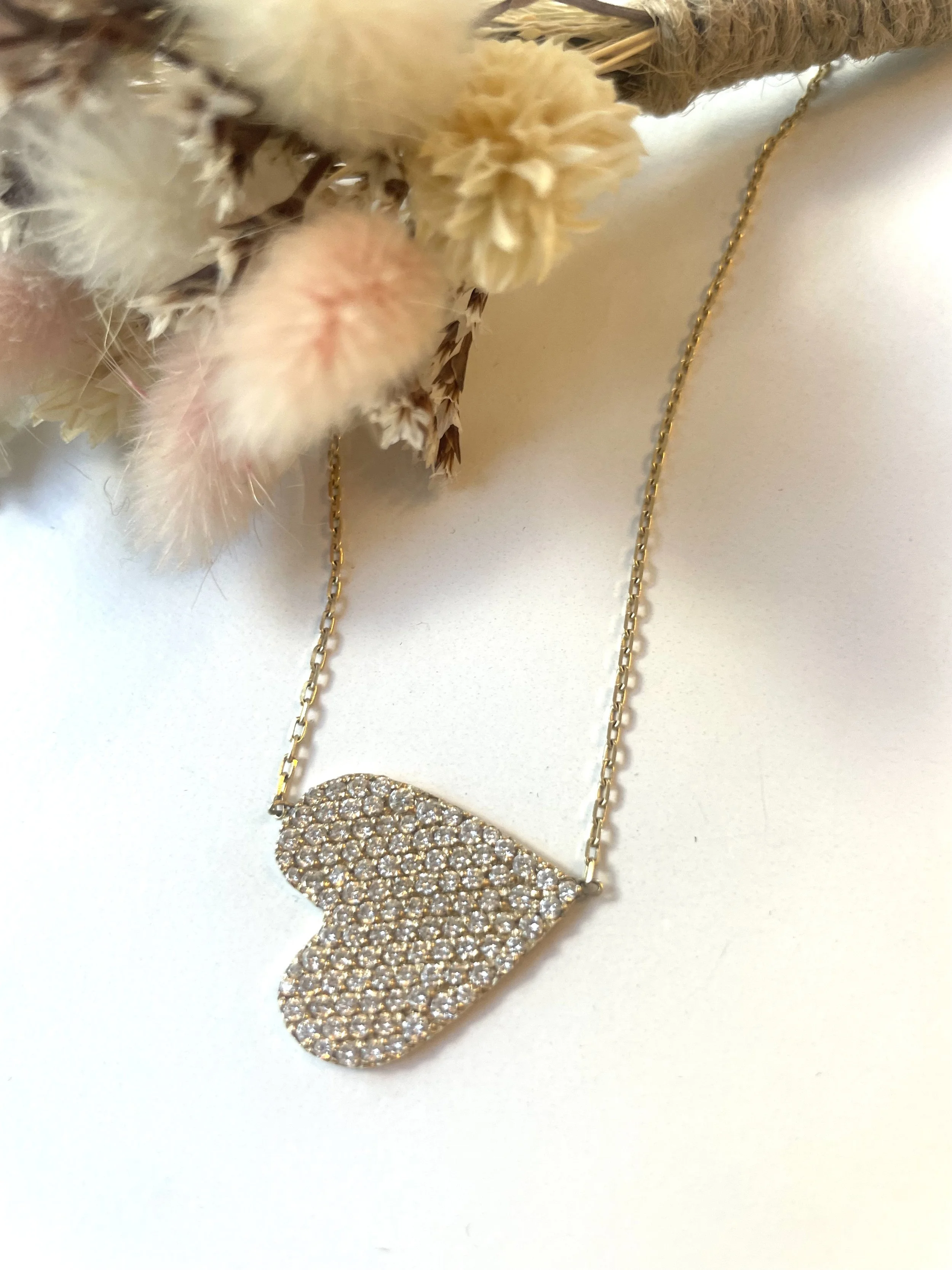 Gold chain necklace with a heart-shaped pendant encrusted with small sparkling stones, hanging near dried flowers on a plain surface.