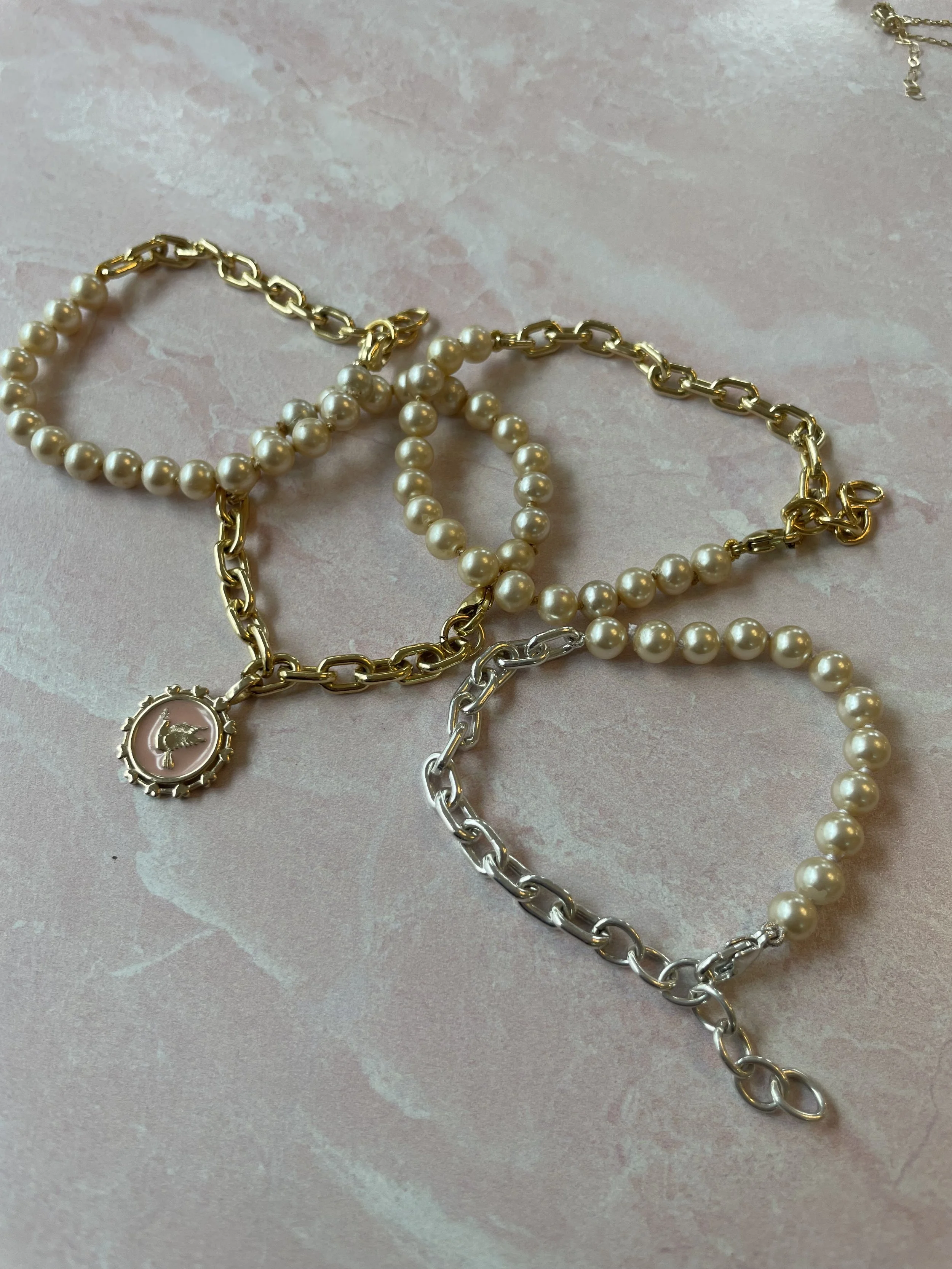 Four bracelets, including two pearl and two chain link, with one pearl bracelet featuring a pendant with a bird design, laid on a pink textured surface.