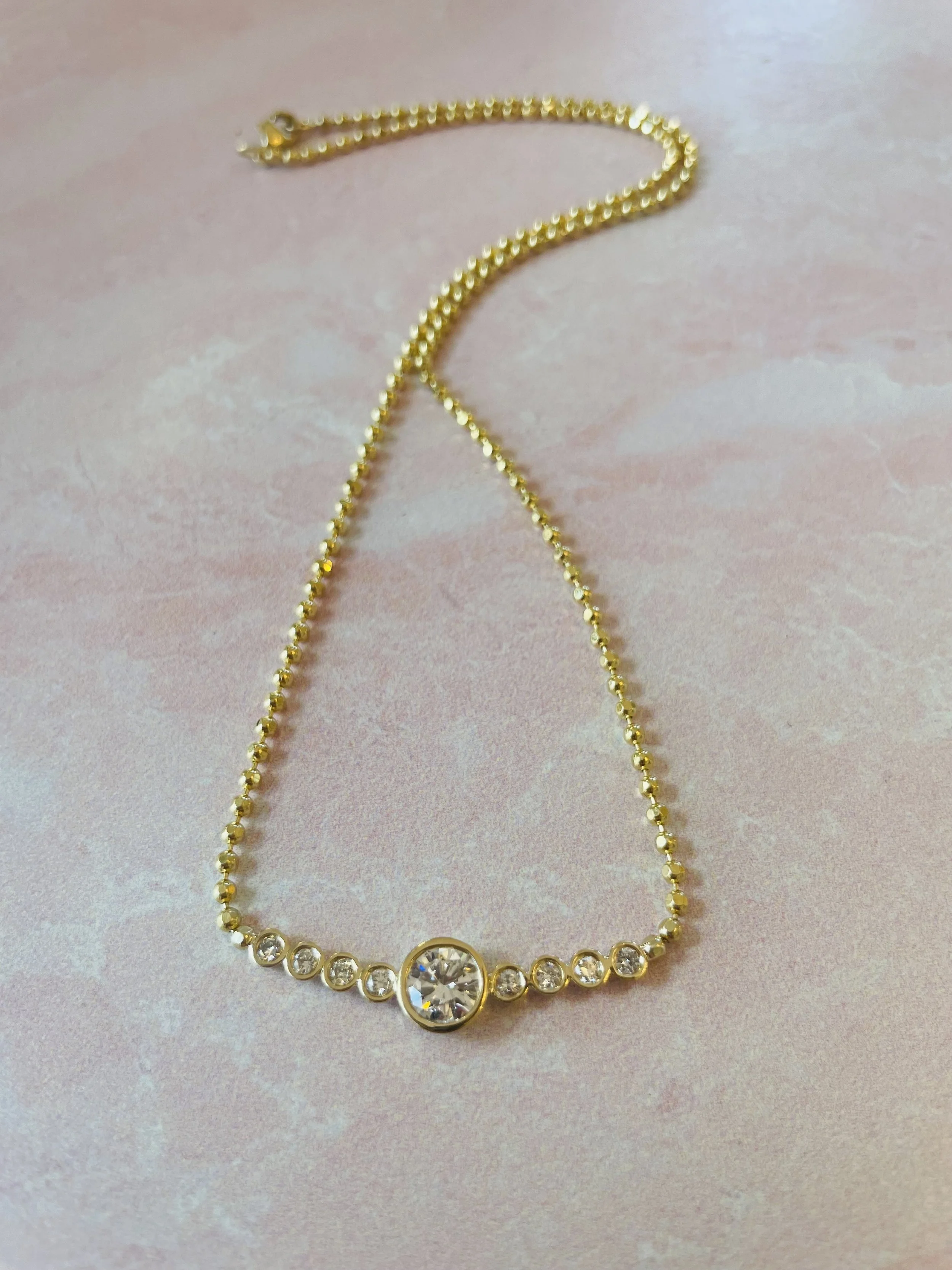 Gold necklace with a central large clear gemstone surrounded by smaller clear gemstones, laid on a pink marble surface.