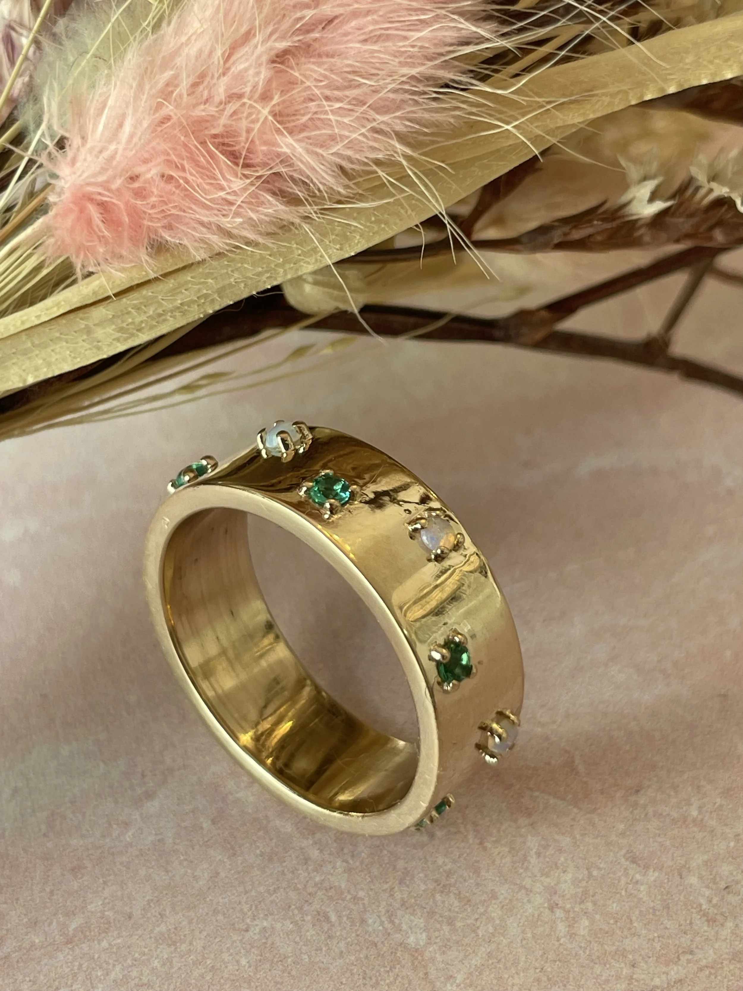 Gold ring with small green and clear gemstones, placed on a pink surface near dried pink and beige flowers.