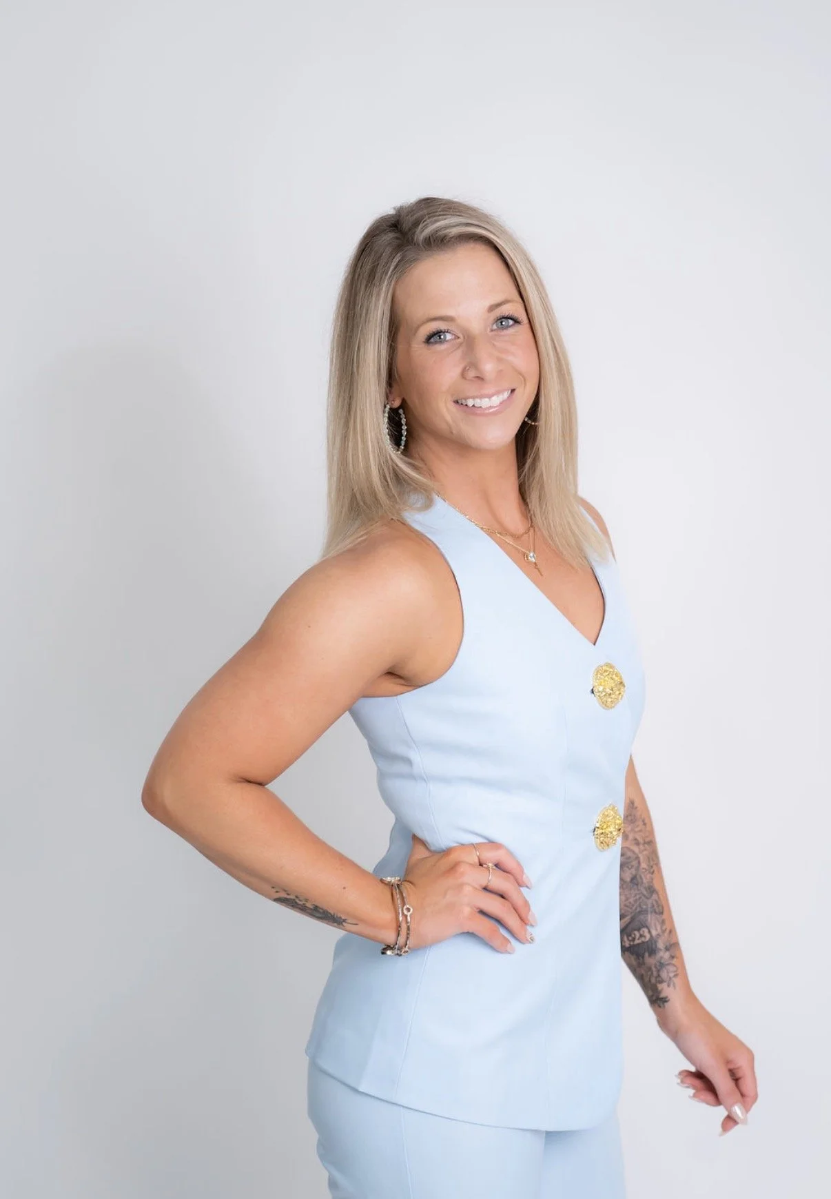 A woman with blonde hair, wearing a light blue sleeveless dress with gold buttons, standing with her hand on her hip and smiling, against a plain light grey background.