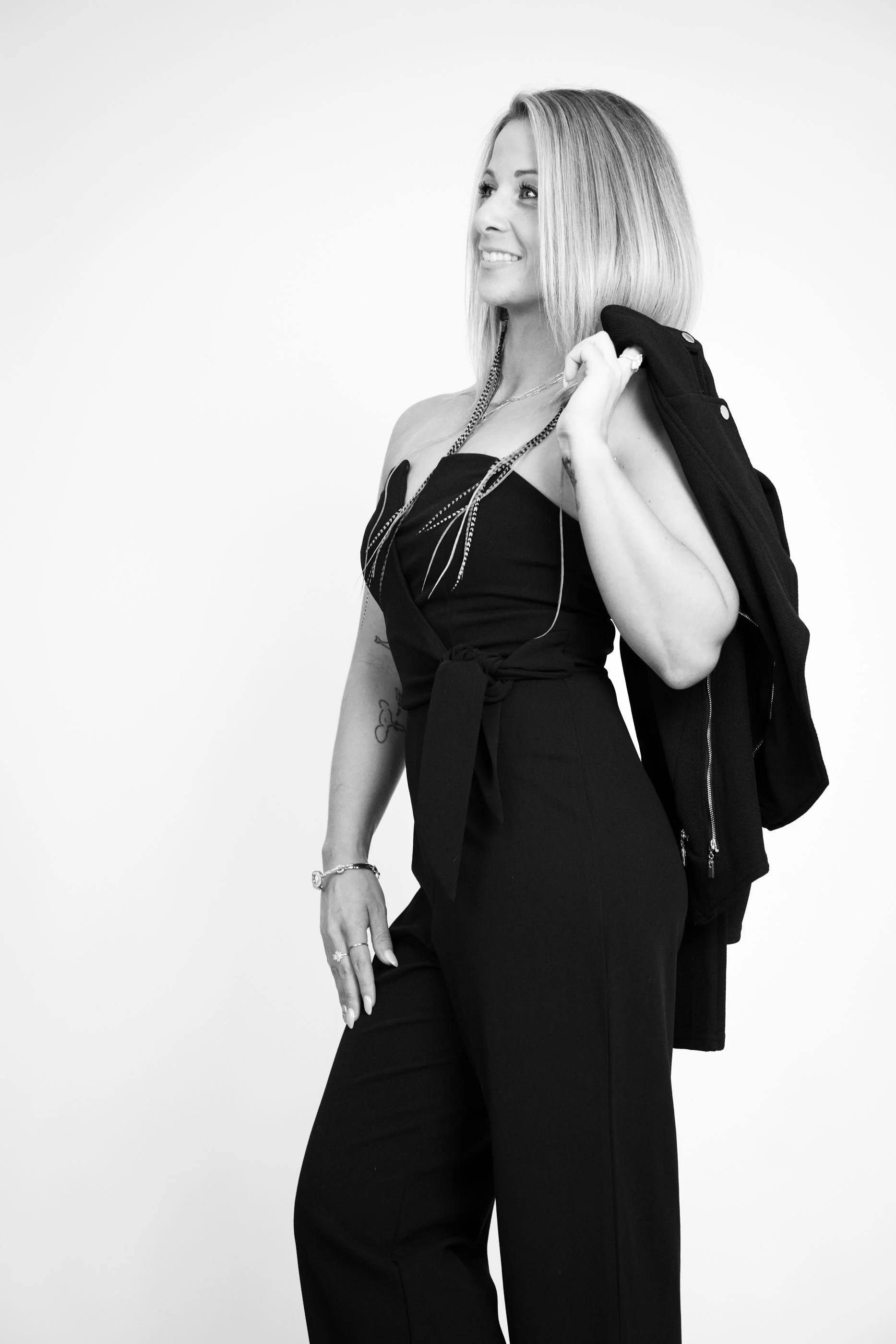 A woman with blonde hair wearing a black strapless jumpsuit, holding a jacket over her shoulder, and smiling while looking to the side.