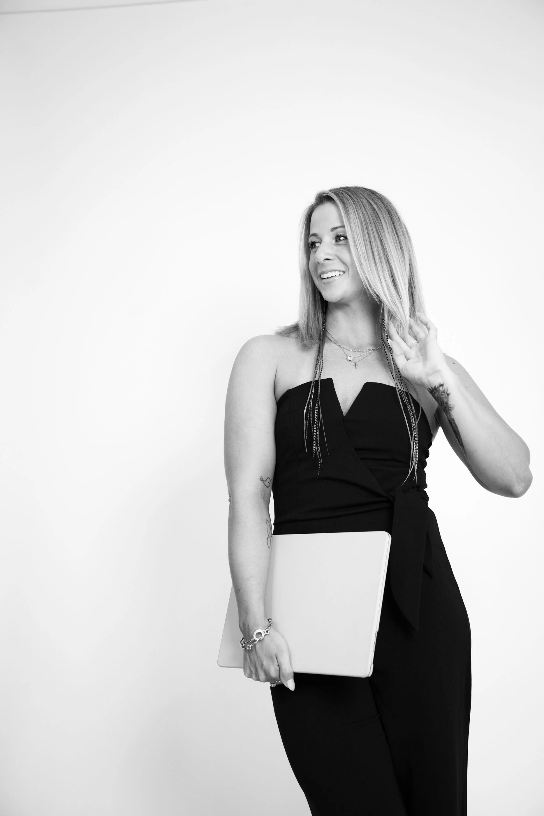 Black and white photo of a woman holding a laptop, wearing a strapless black jumpsuit, with tattoos on her arms, and smiling while looking to the side.