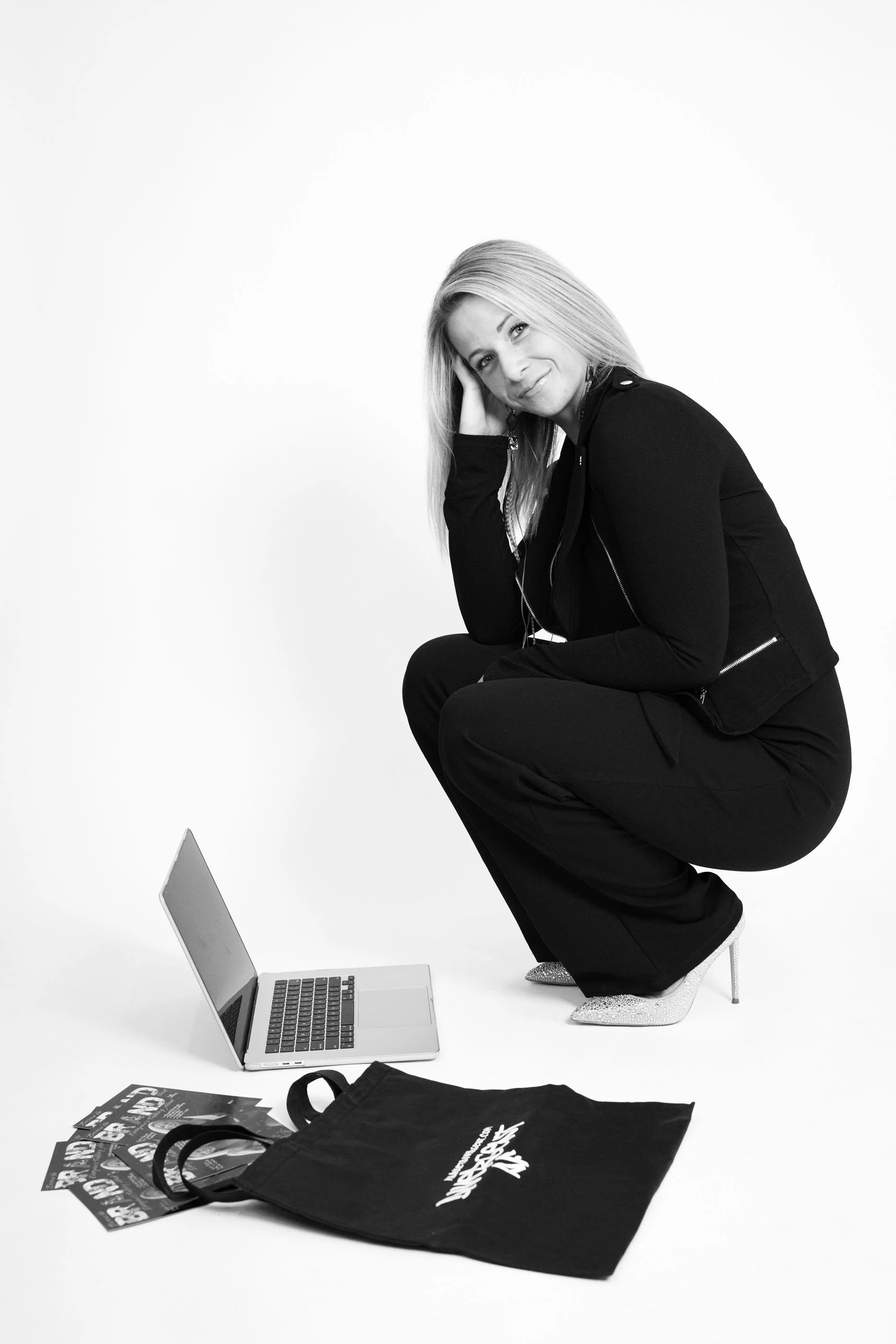 A woman with long blonde hair crouching on her heels, wearing a black jacket and pants, silver high heels, and earbuds, smiling at the camera. There is a laptop, some flyers, and a black tote bag on the floor in front of her, all set against a plain white background.
