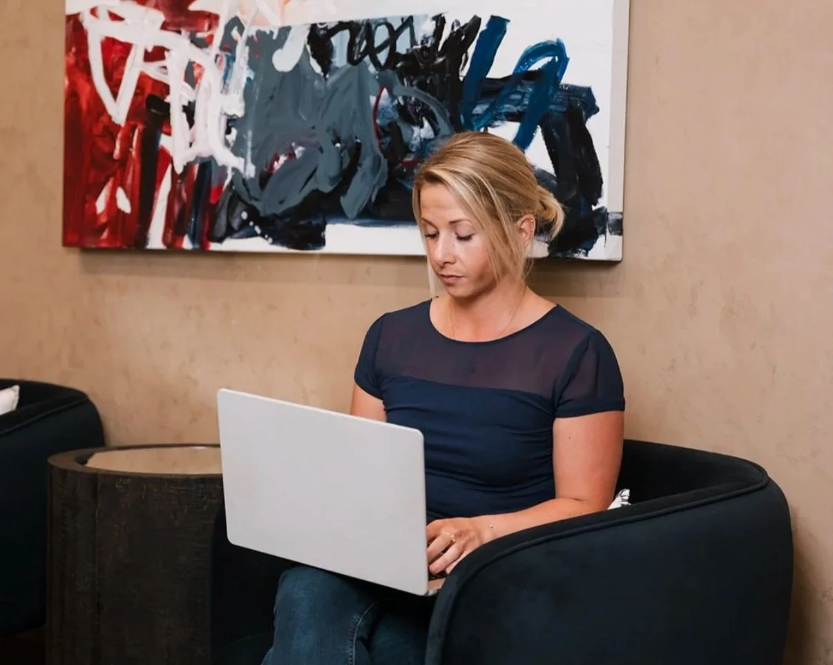 A young woman with blonde hair tied back sitting on a black chair, working on a silver laptop. She is wearing a navy blue top with sheer fabric on the shoulders. Behind her, there is a beige wall with an abstract painting featuring red, white, black, and blue strokes.