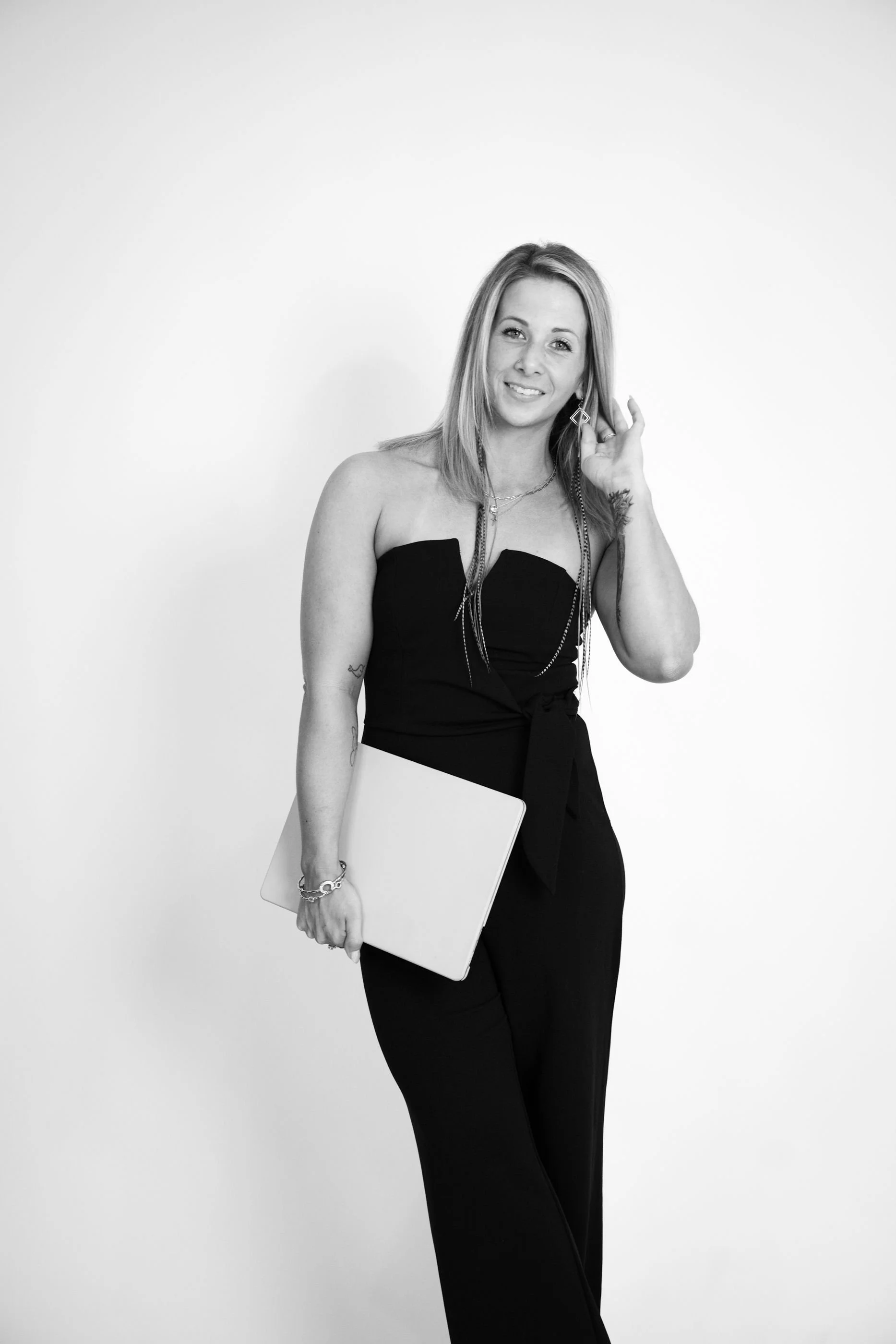 Black-and-white portrait of a woman with long hair, smiling, holding a laptop, wearing a strapless black outfit and jewelry, standing against a plain background.